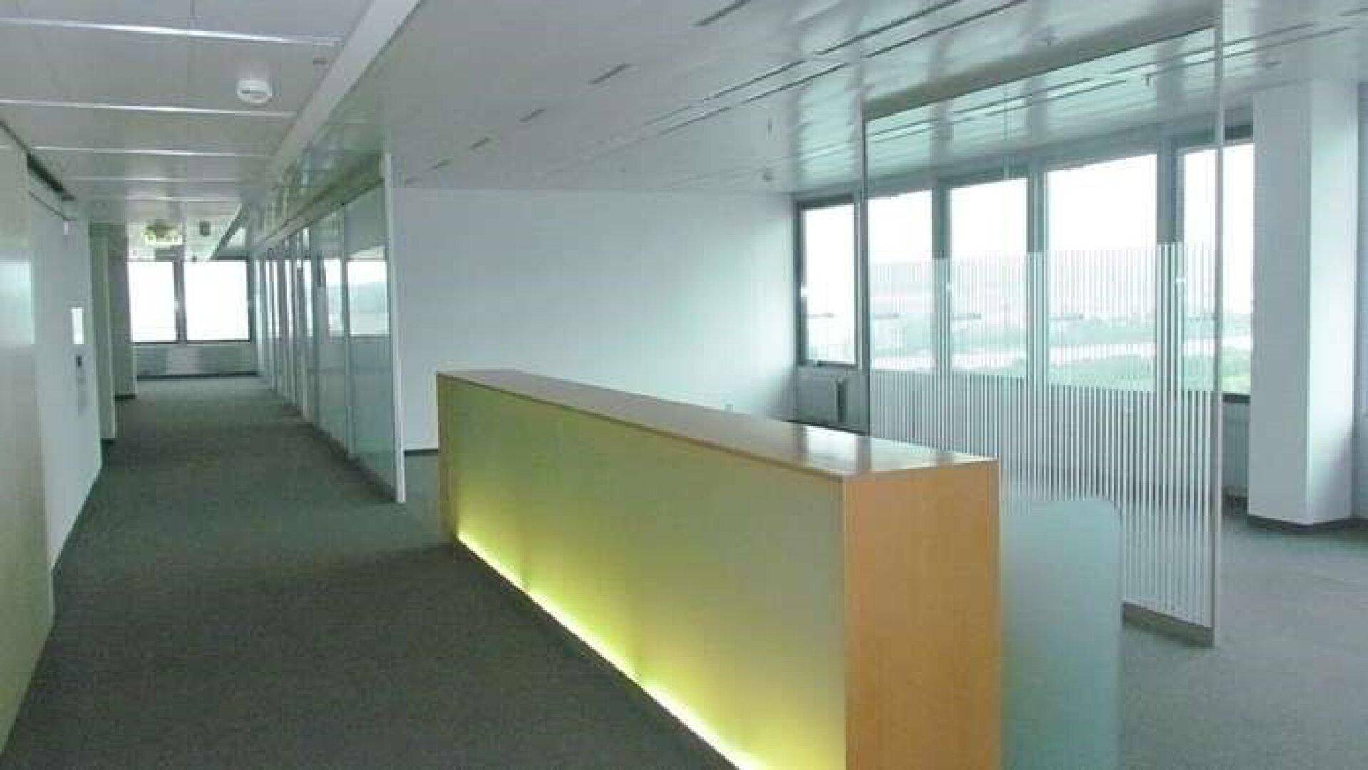 Heller Bürobereich mit Glaswänden, Teppichboden und einer beleuchteten Empfangstheke.