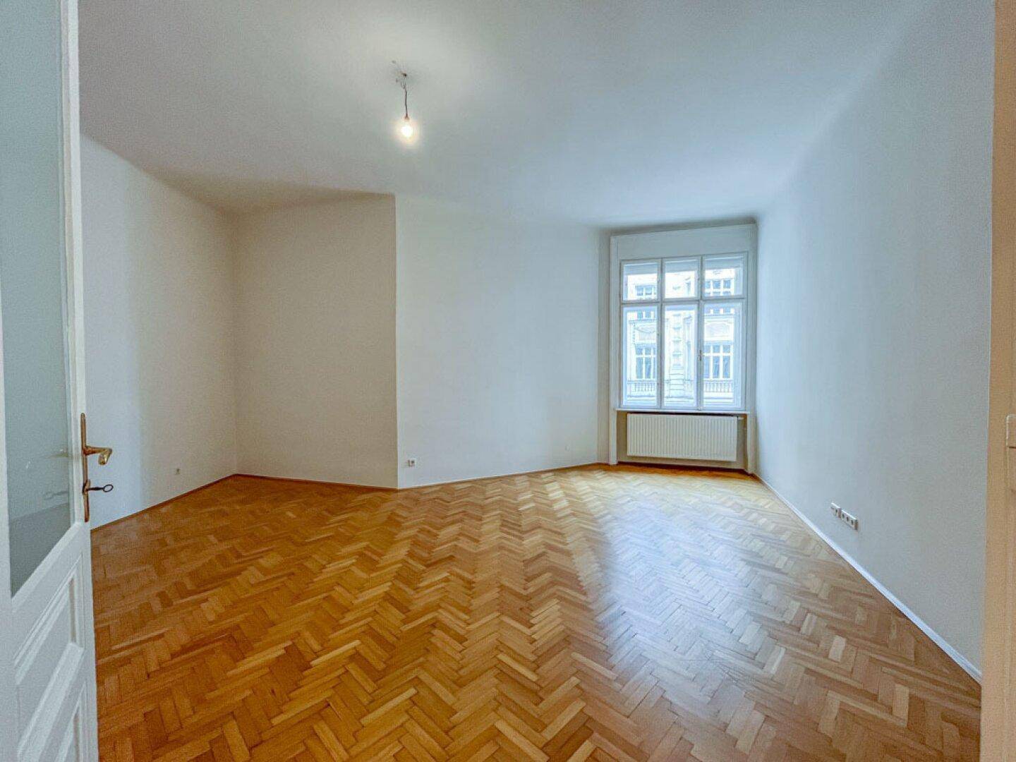 Helles, leeres Zimmer mit klassischem Fischgrätparkett und einem großen Fenster, das viel Licht hereinlässt.