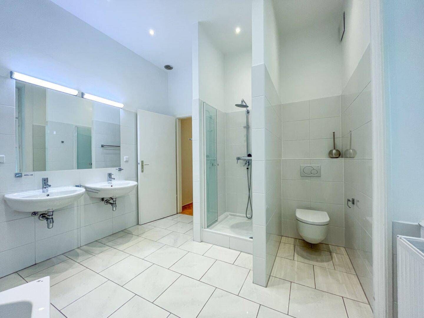 Modernes Badezimmer mit weißen Fliesen, Doppelwaschbecken, einer begehbaren Dusche und Wand-WC.