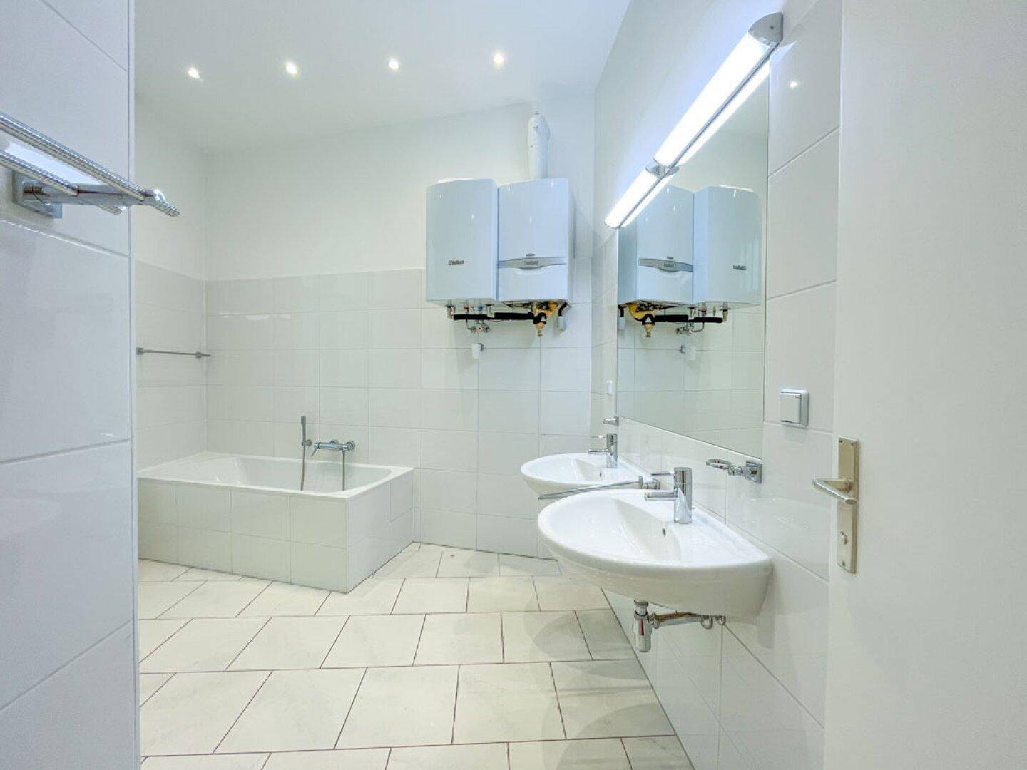 Helles Badezimmer mit weißen Fliesen, einer Badewanne, Doppelwaschbecken und zwei Warmwasserbereitern.