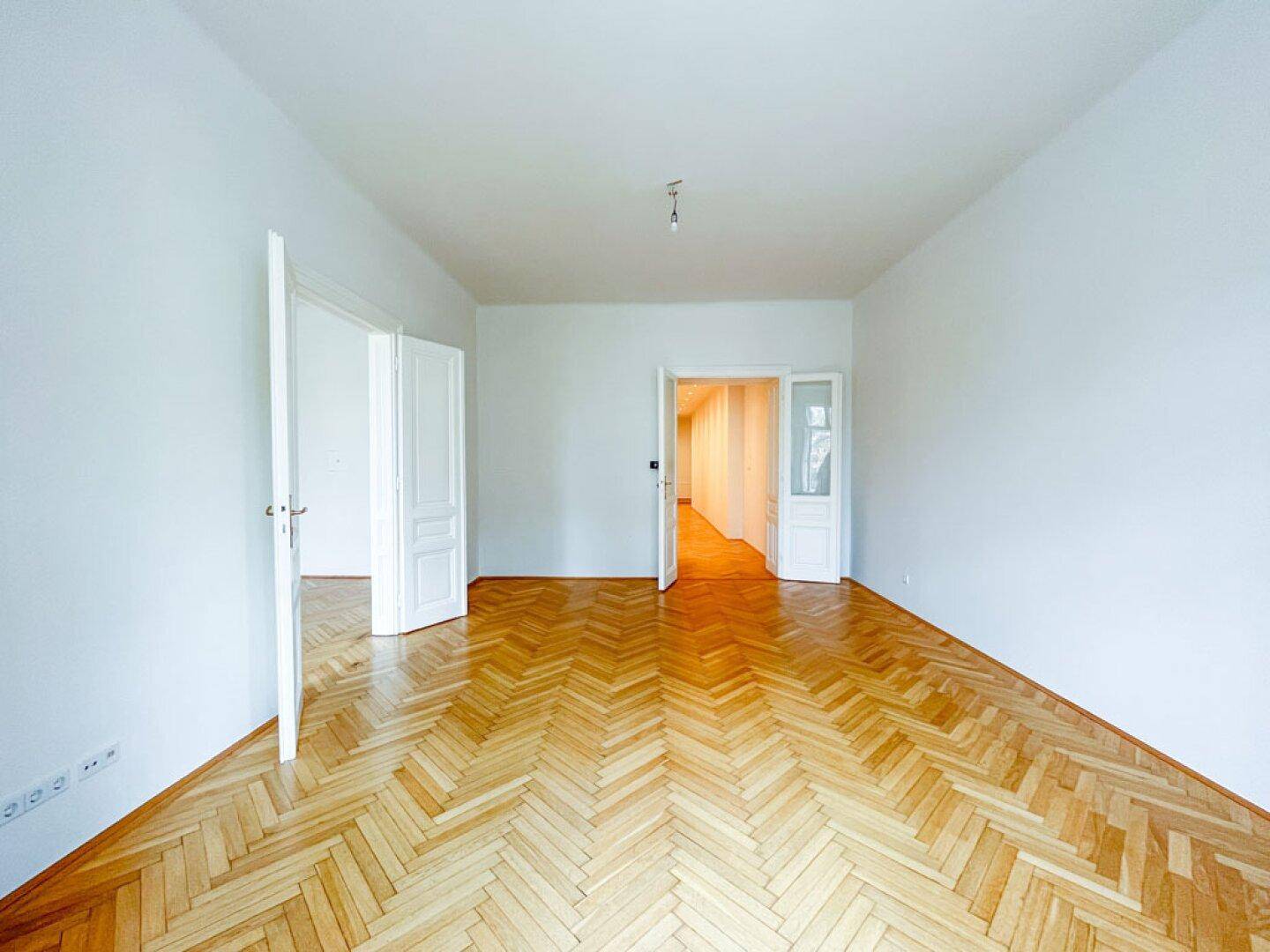 Leeres Zimmer mit elegantem Fischgrätparkett und Zugang zu einem langen Flur mit Einbaustrahlern.