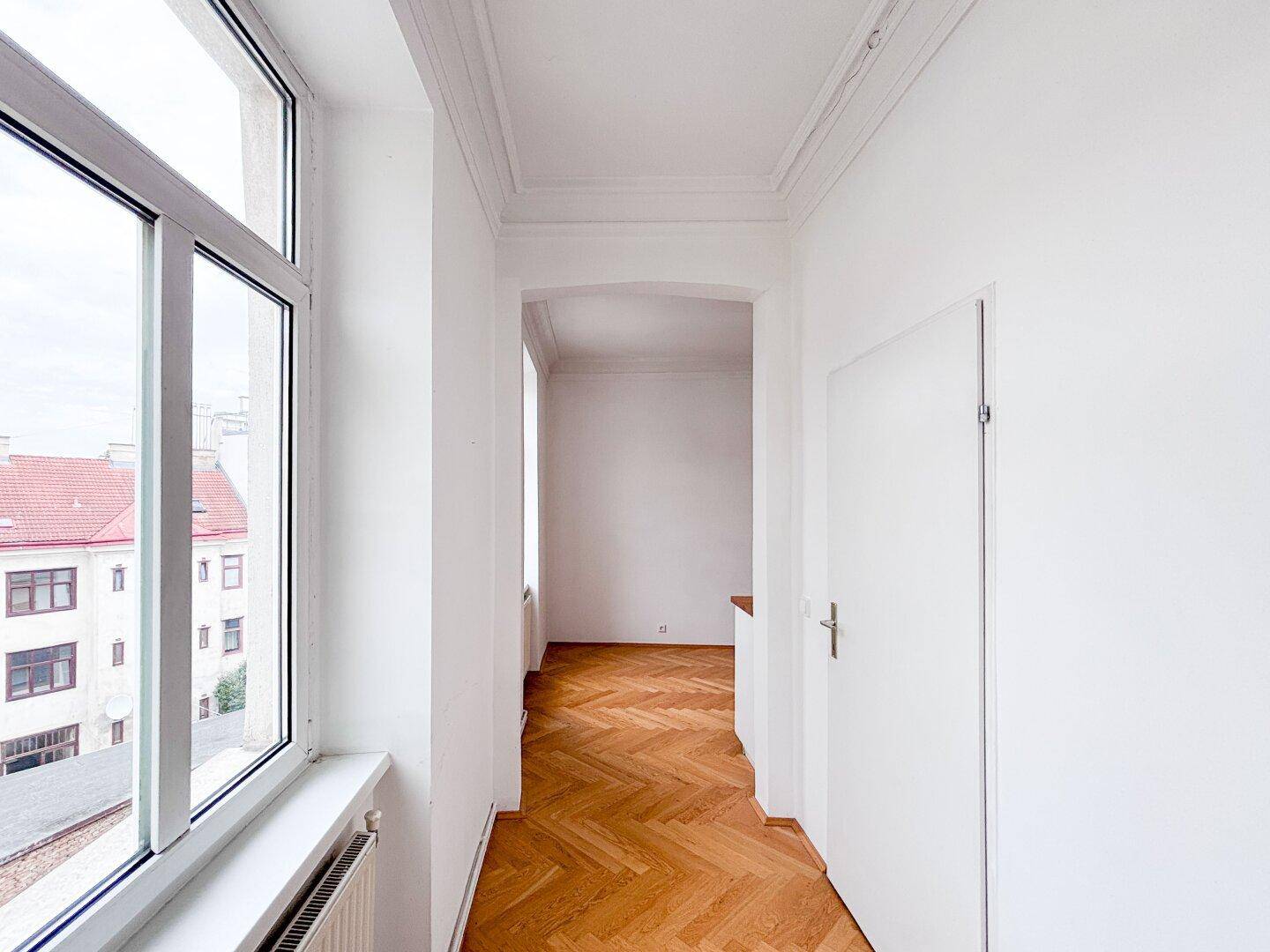 Heller Flur mit Fischgrätparkett und Fenster, das einen Blick auf die Dächer bietet.
