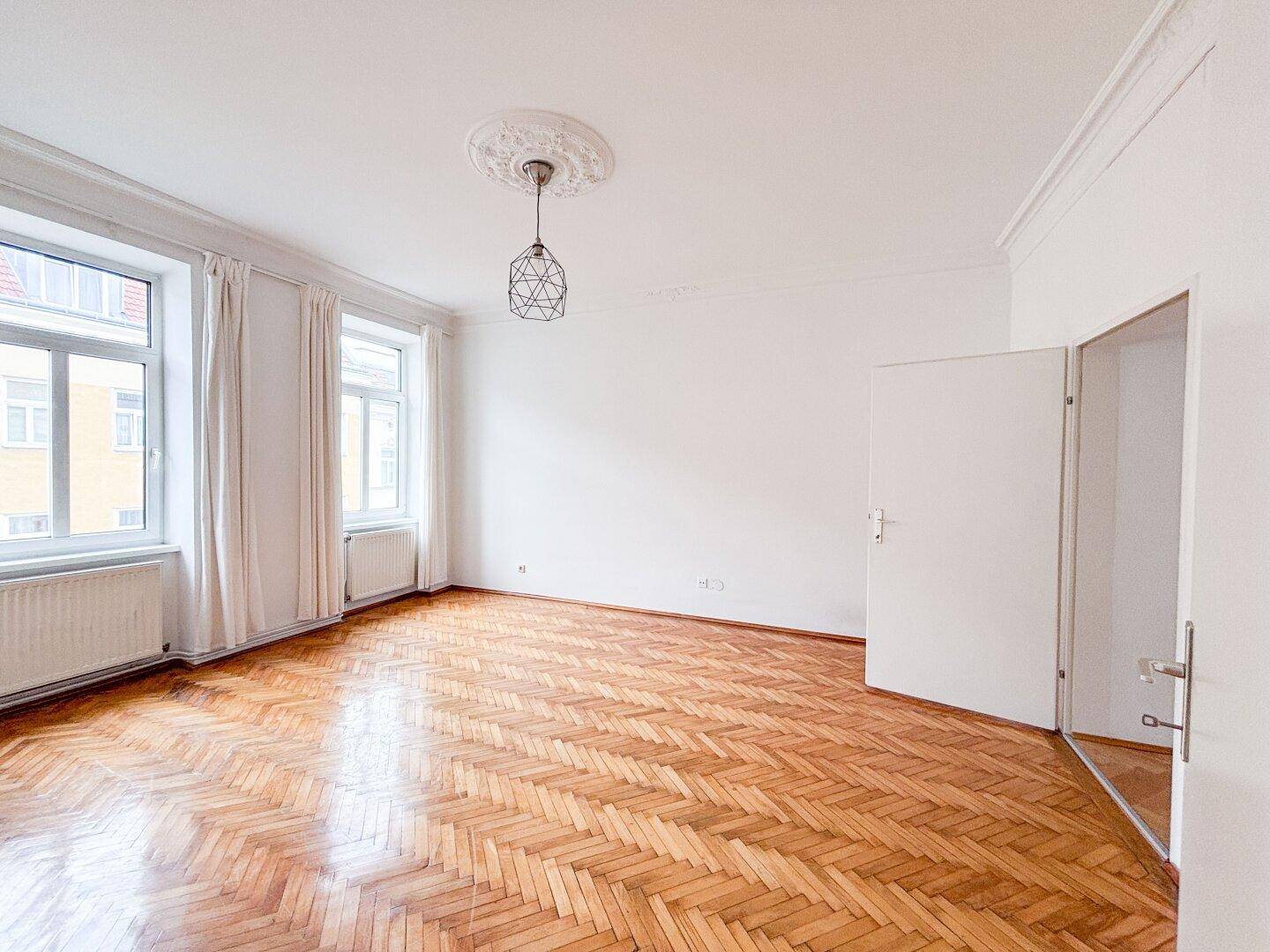 Lichtdurchflutetes Wohnzimmer mit klassischem Fischgrätparkett und hohen Decken, bereit zur Einrichtung.