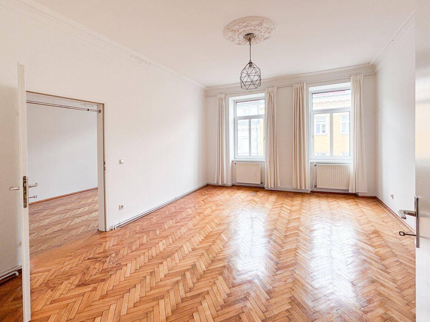 Geräumiges, leeres Wohnzimmer mit Fischgrätparkett, Stuckdecke und großen Fenstern.
