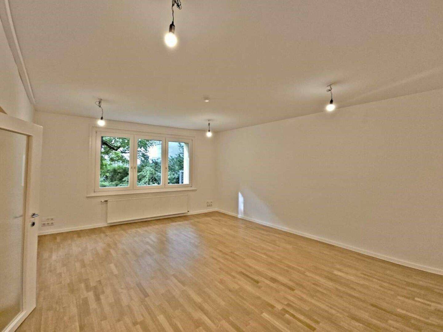 Heller, leerer Raum mit großen Fenstern, Holzboden und Blick ins Grüne.