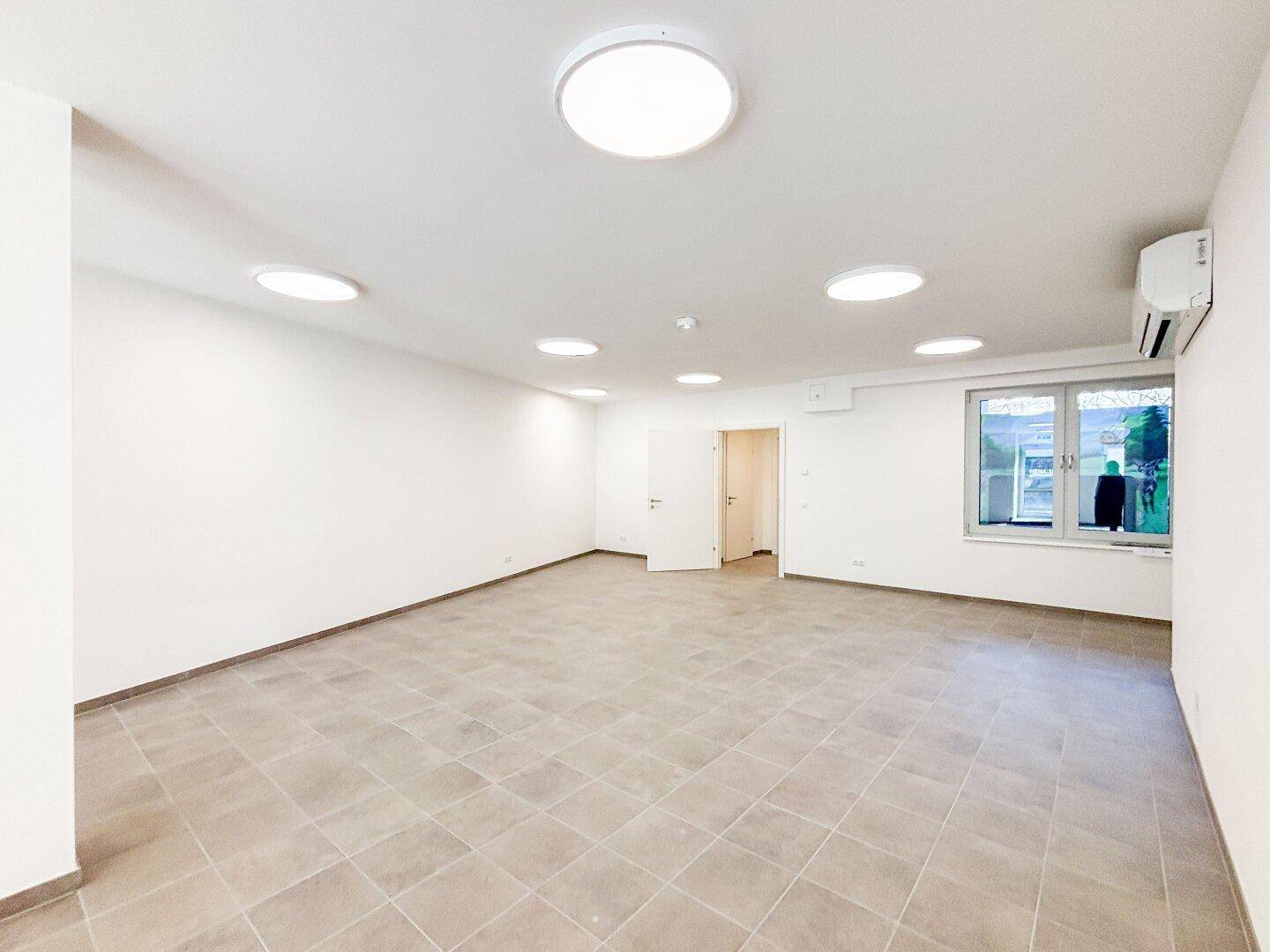 Geräumiger, leerer Gewerberaum mit Fliesenboden, Fenster und Klimaanlage, ideal für Büros oder Praxen.