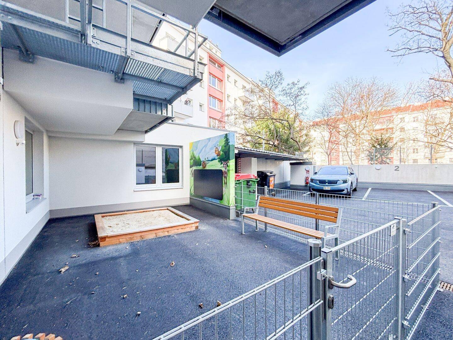 Außenbereich mit Sandkasten, Sitzbank und angrenzendem Parkplatz, umgeben von Wohngebäuden.