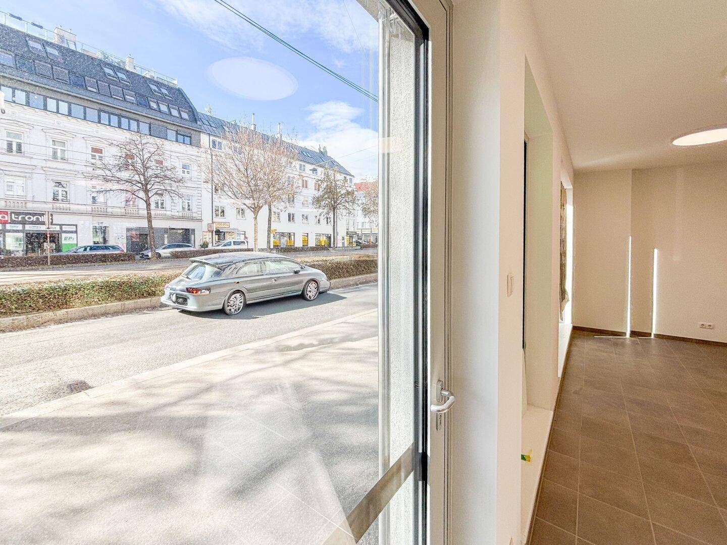 Blick aus dem Fenster eines Gewerberaums auf eine belebte Stadtstraße mit Gebäuden und Fahrzeugen.