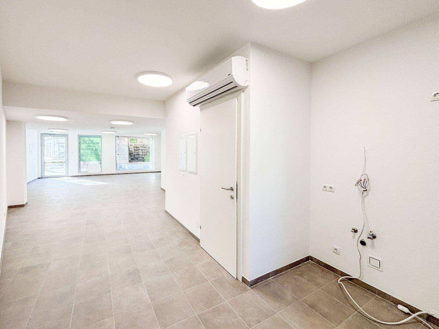 Geräumiger, leerer Gewerberaum mit Fliesenboden, Klimaanlage und Zugang zu weiteren Bereichen, ideal für flexible Nutzung.
