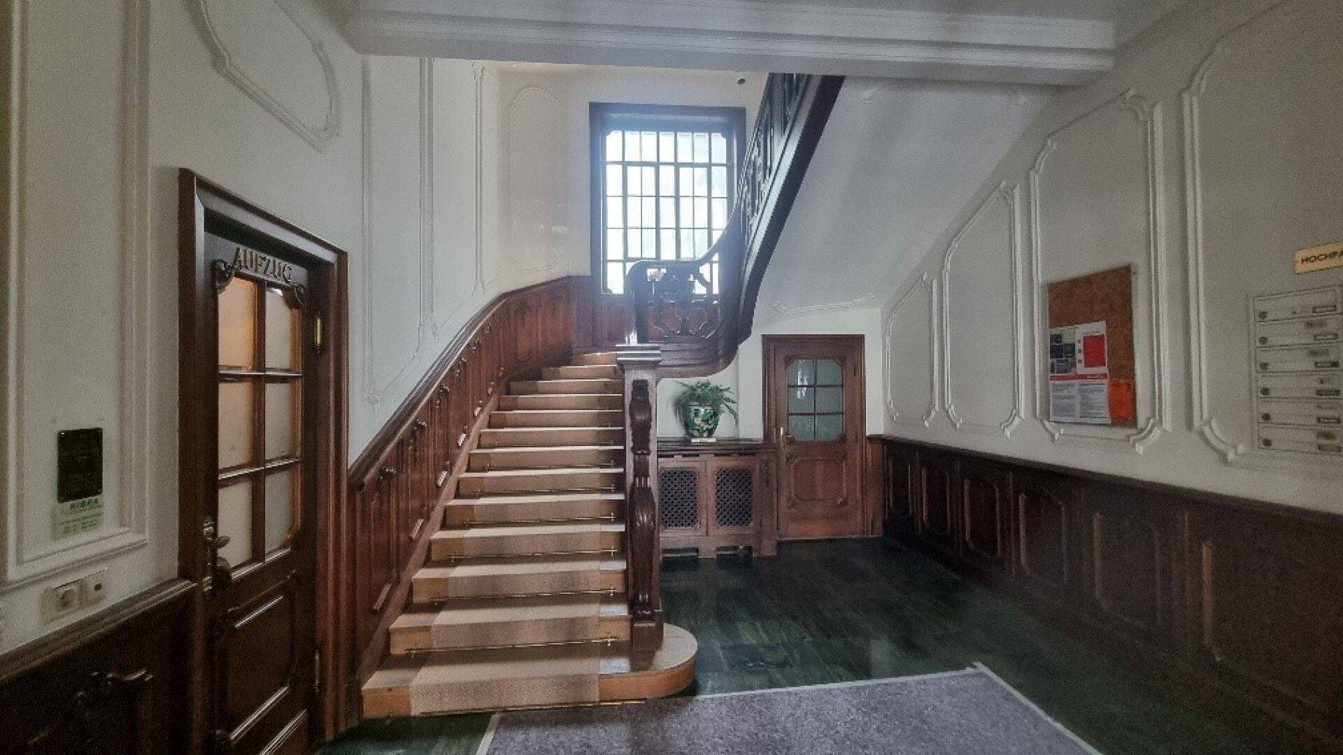 Elegantes Treppenhaus mit Holzgeländer, Marmorstufen und Zugang zu einem Aufzug in einem Altbau.