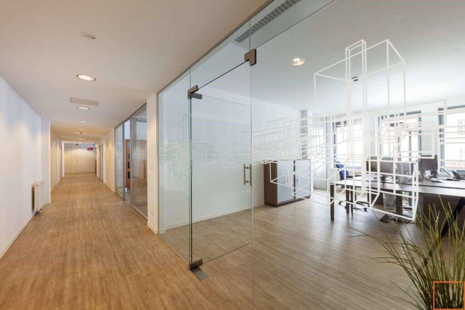 Langer Büroflur mit modernen Glaswänden, die Einblicke in die Büros gewähren, und hellem Holzboden.