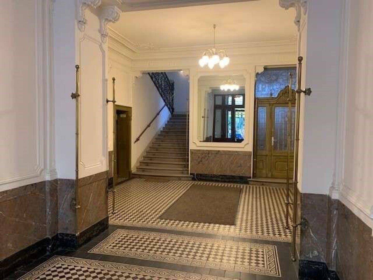 Geräumiger Eingangsbereich eines Altbaus mit Schachbrettmuster-Fliesenboden und eleganter Treppe.