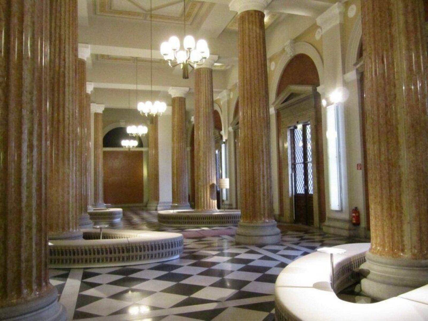 Prunkvoller Flur mit hohen Säulen, Schachbrettmuster-Boden und opulenten Kronleuchtern in einem historischen Gebäude.