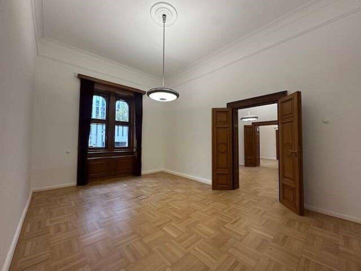 Geräumiger, heller Raum mit klassischem Fischgrätparkett, hohen Decken und einem großen Fenster mit dunklen Vorhängen.