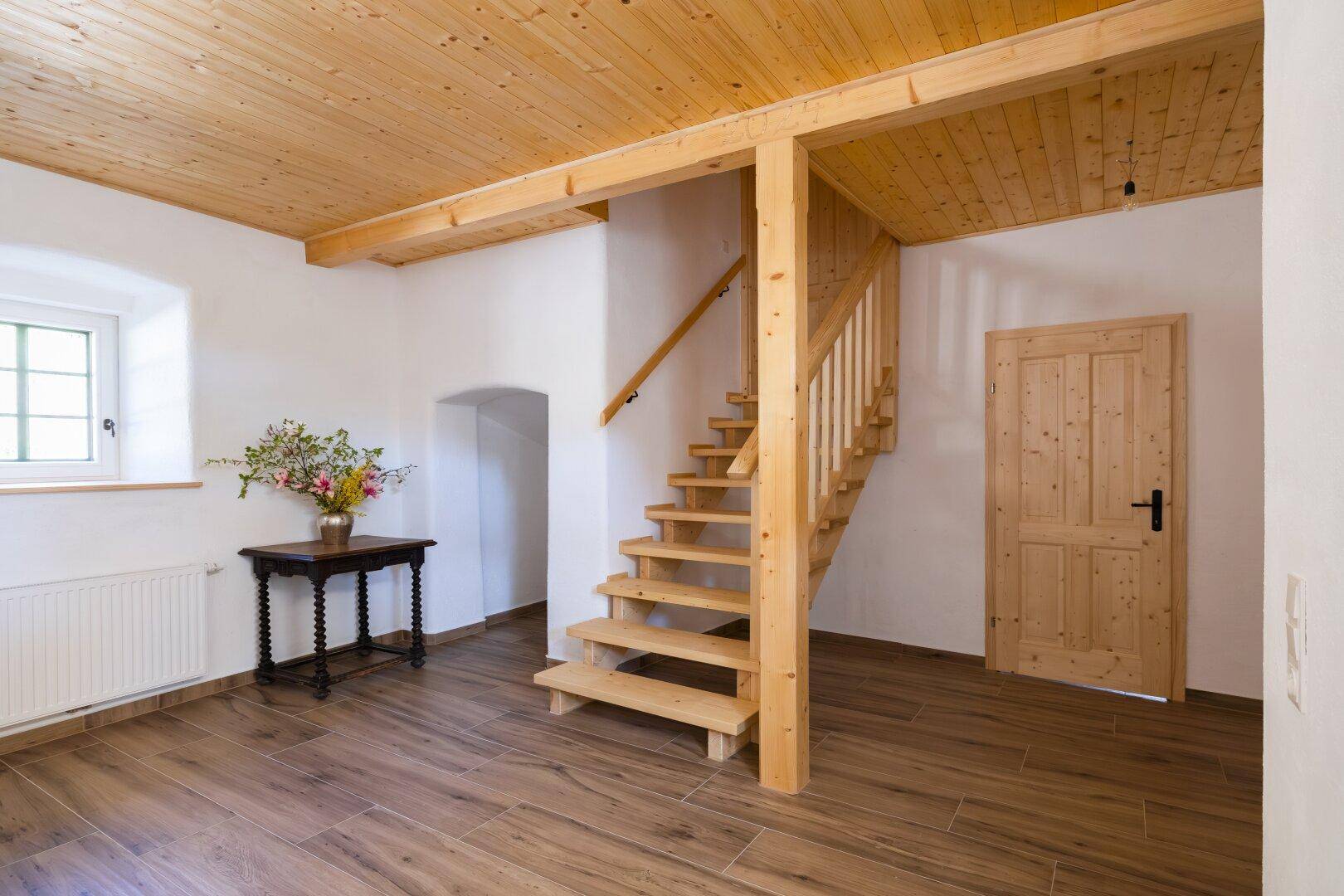 Heller Flur mit rustikaler Holztreppe, Holzbalkendecke und modernen Fliesenboden.