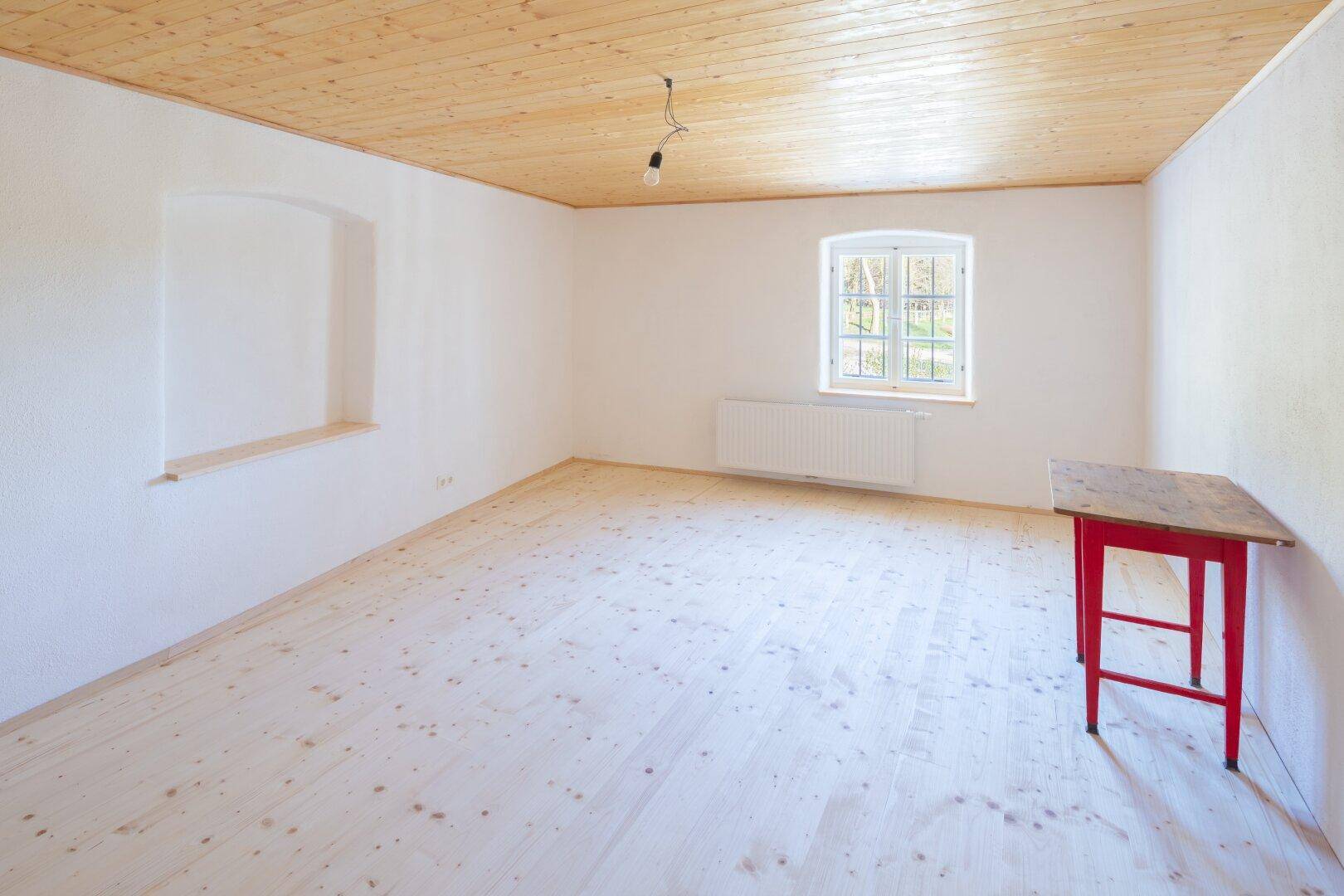 Helles, leeres Zimmer mit hellem Holzfußboden, weiß gestrichenen Wänden und einem Fenster.