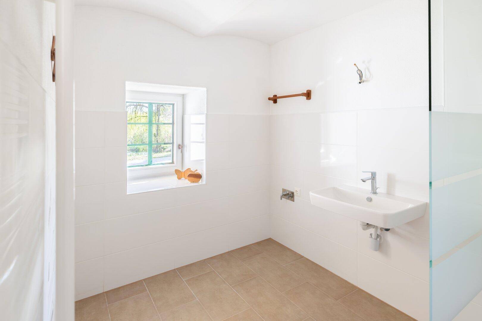 Helles Badezimmer mit weißen Fliesen, Waschbecken und einem Fenster, sauber und funktional.