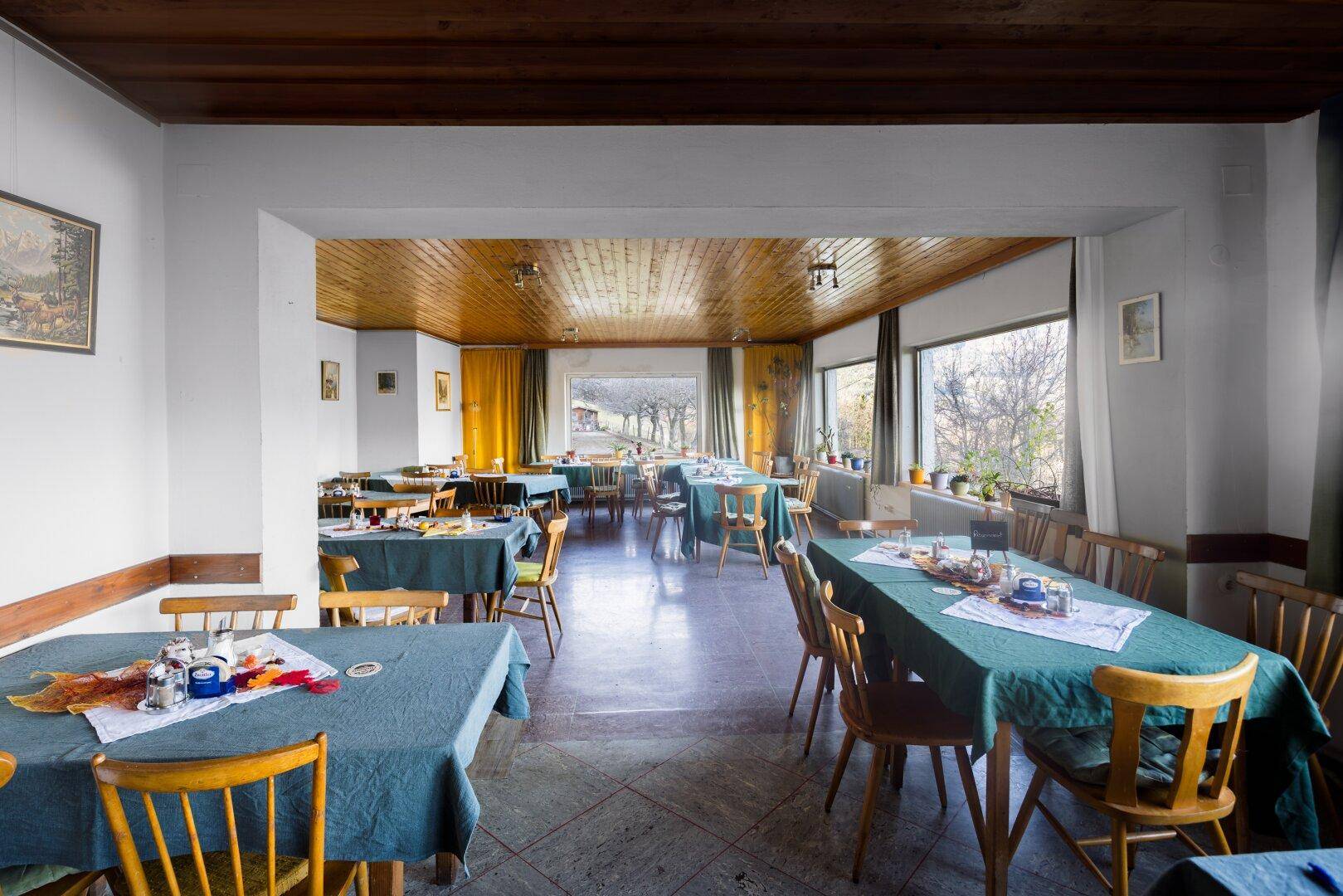 Geräumiger Speisesaal mit mehreren Tischen, Holzdecken und großen Fenstern mit Bergblick.