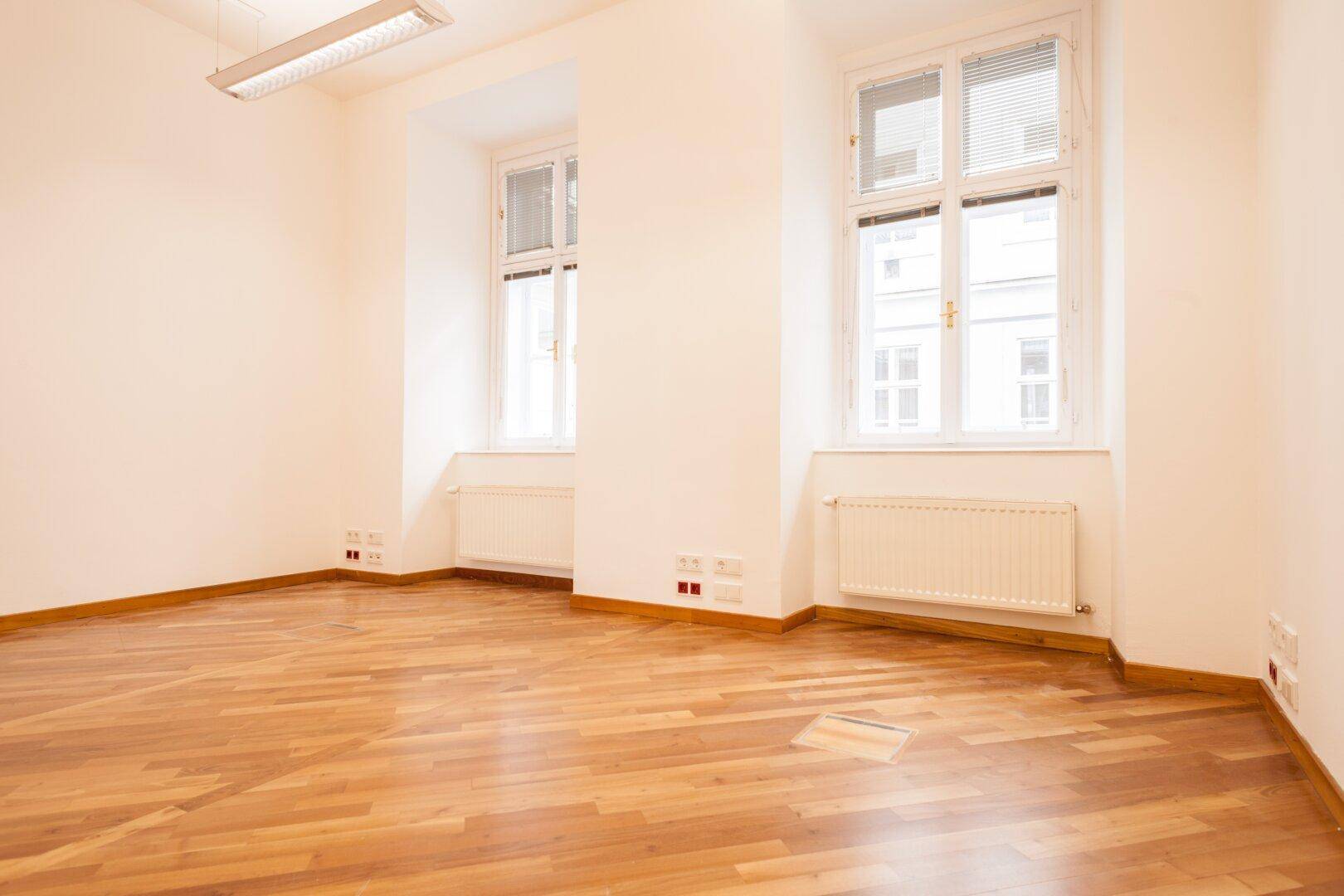 Heller, leerer Raum mit Holzboden und zwei großen Fenstern, die viel Tageslicht hereinlassen.