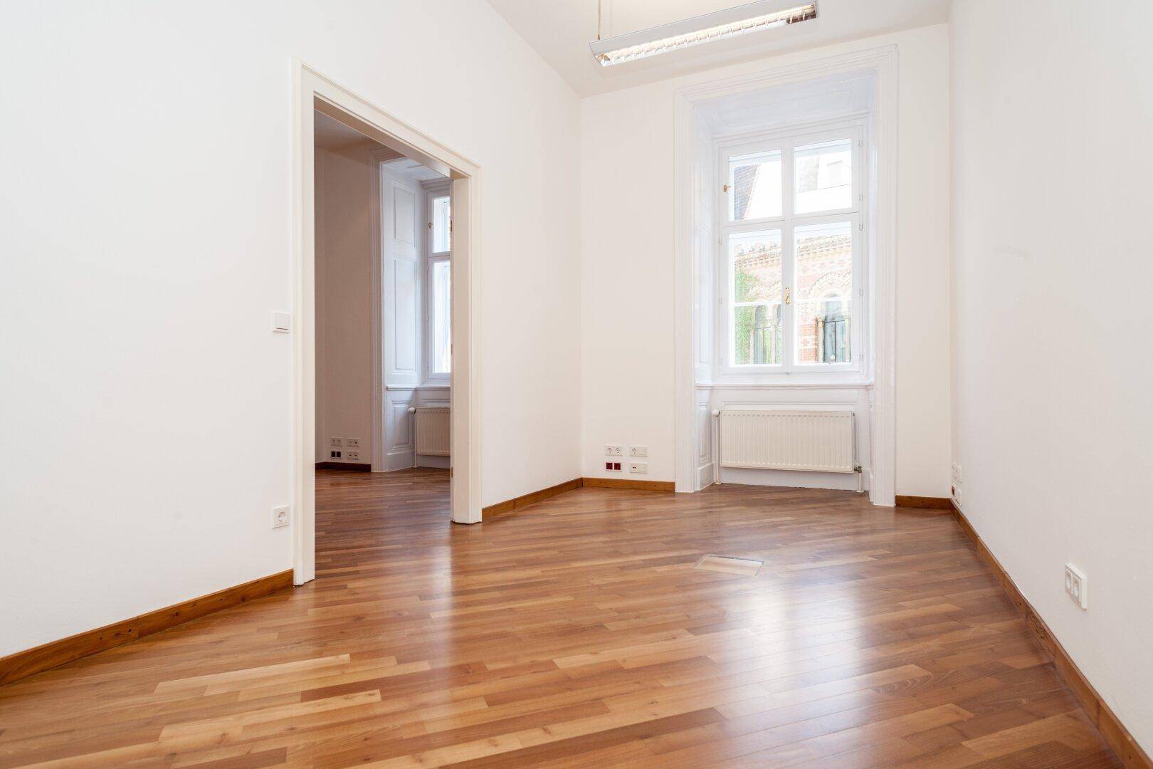 Lichtdurchfluteter Raum mit Holzboden, einem großen Fenster und einem offenen Zugang zu einem Nebenraum.