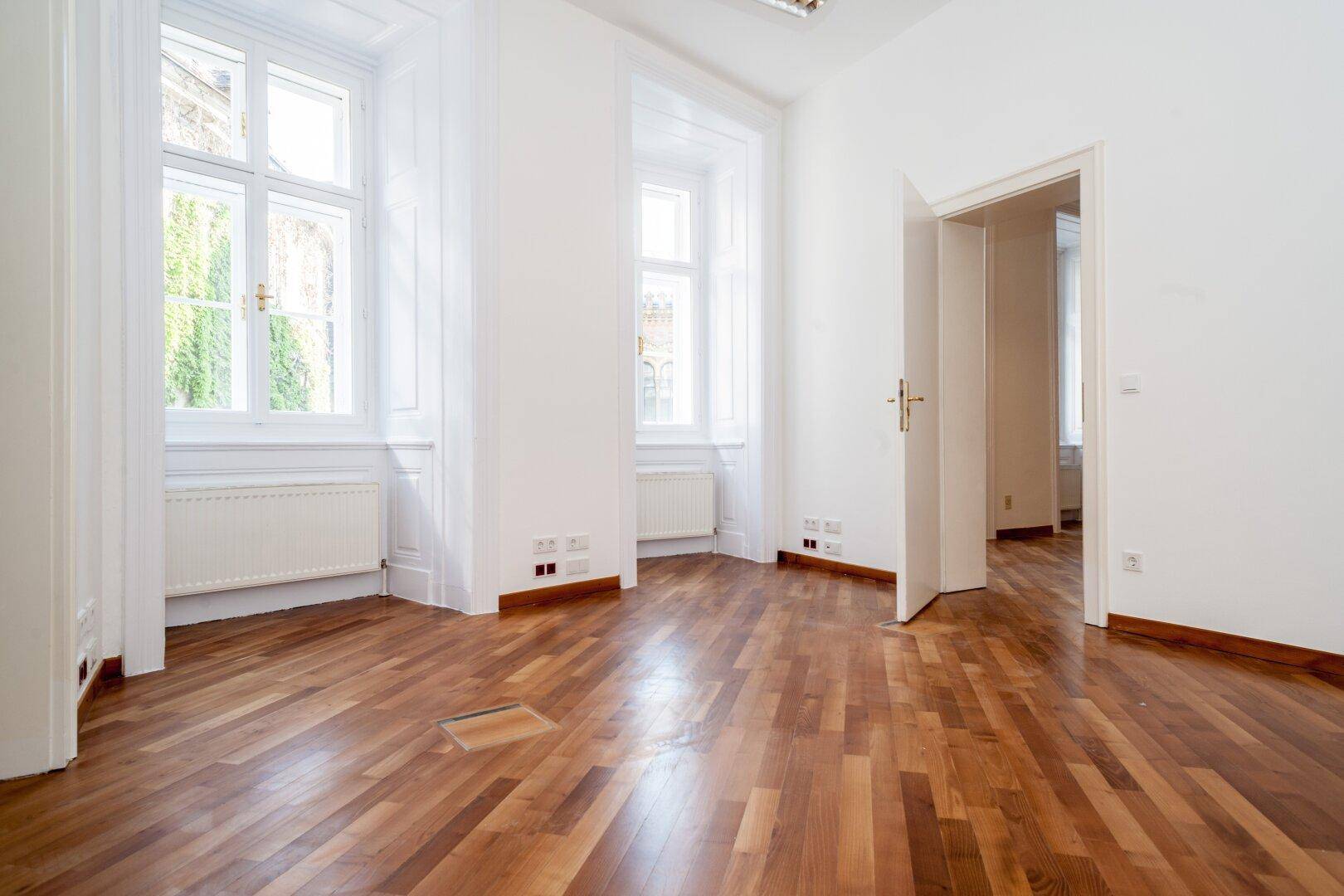Geräumiger, heller Raum mit Holzboden, hohen Fenstern und offenem Durchgang zu einem weiteren Bereich.