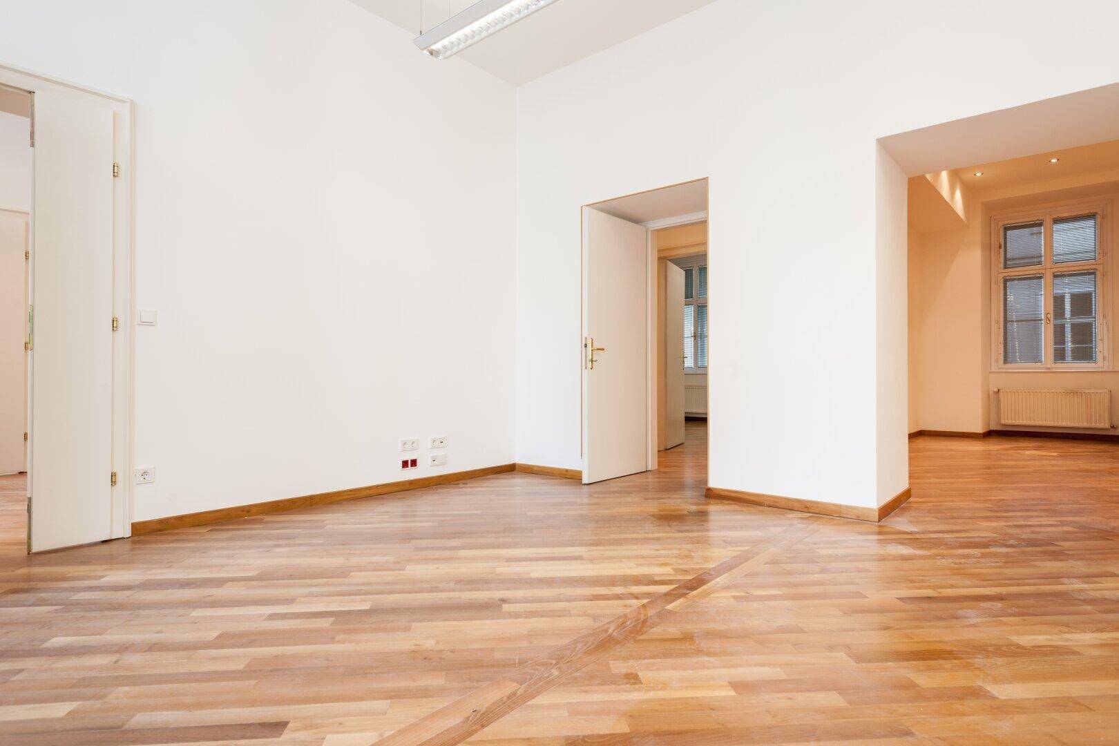 Großer, heller Raum mit Holzboden und mehreren offenen Türen, die zu anderen Zimmern führen.