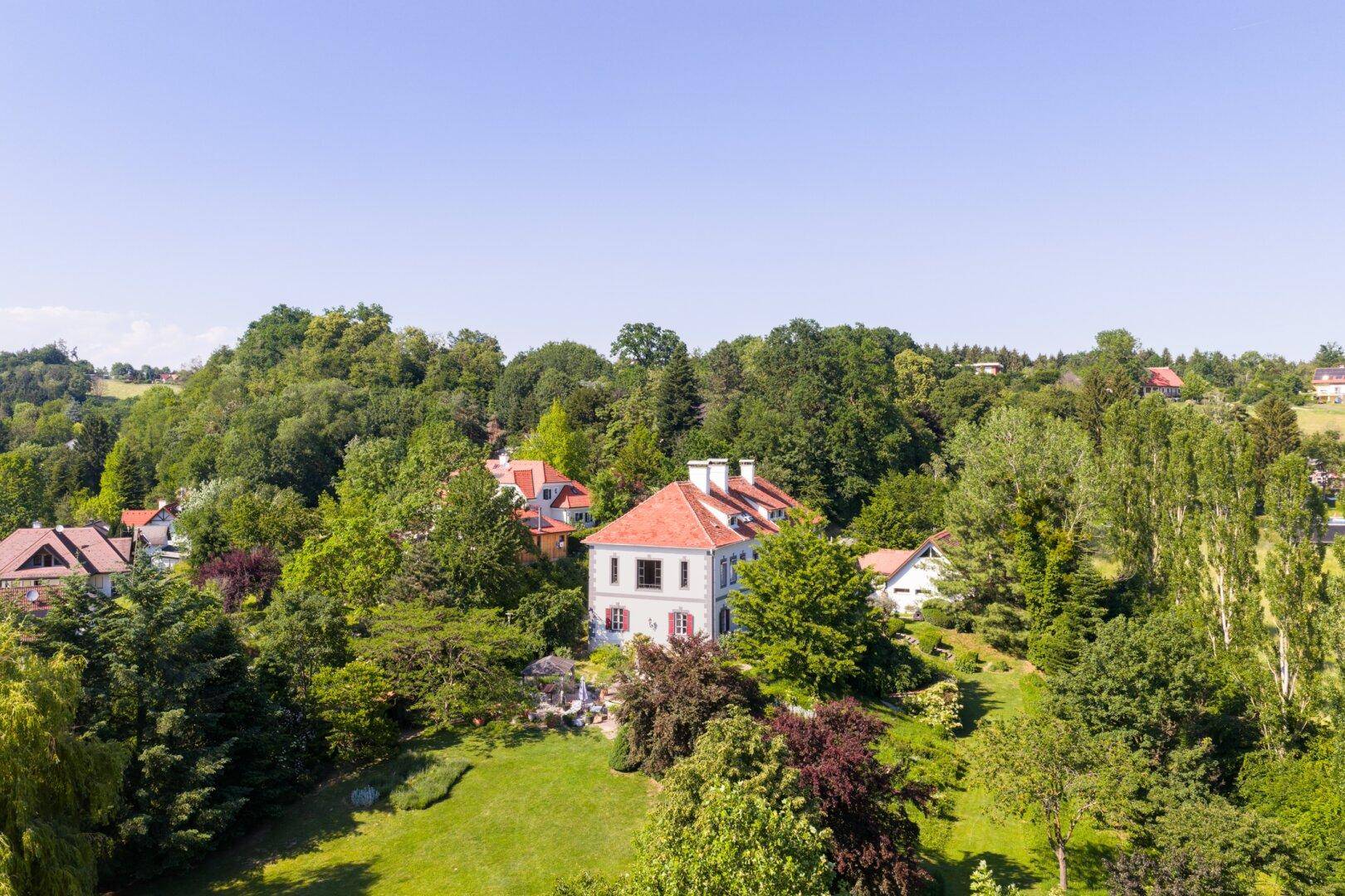 Panoramablick auf die Villa, eingebettet in eine grüne Hügellandschaft mit vielen Bäumen.