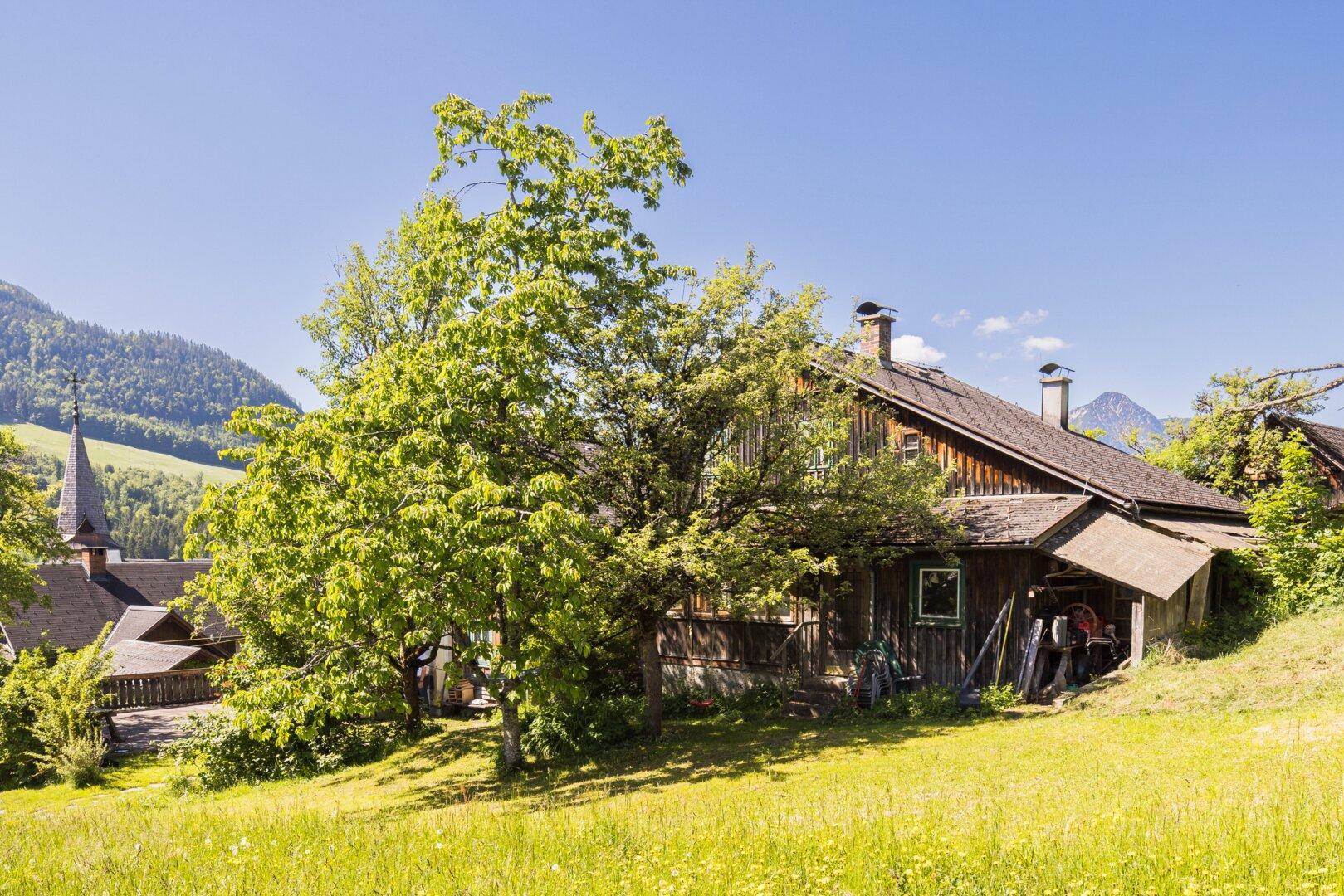 Traditionelles Holzhaus mit Anbau und Blick auf die Kirche in ländlicher Umgebung.