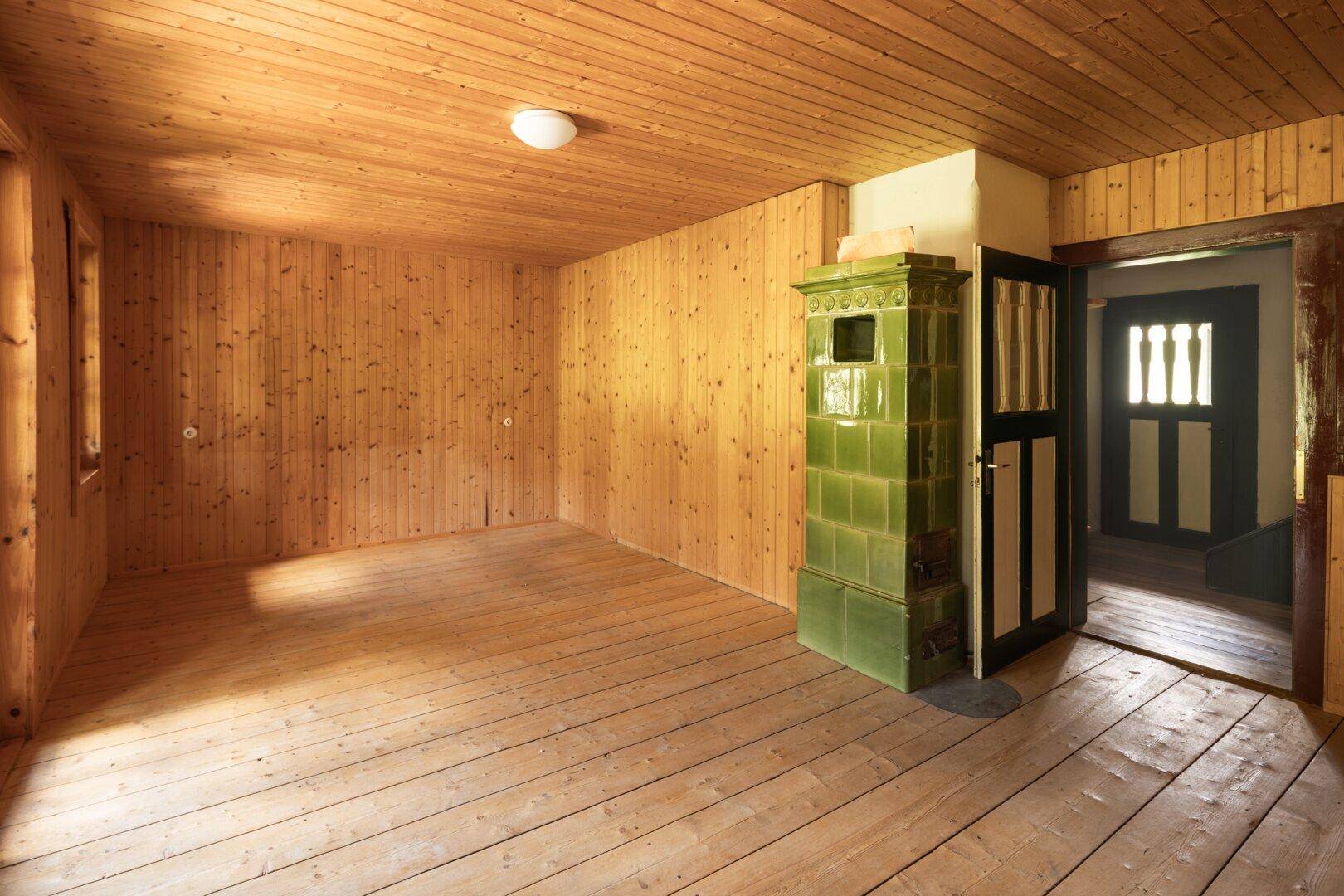 Geräumiger Wohnraum mit Holztäfelung, Holzdielenboden und einem grünen Kachelofen.