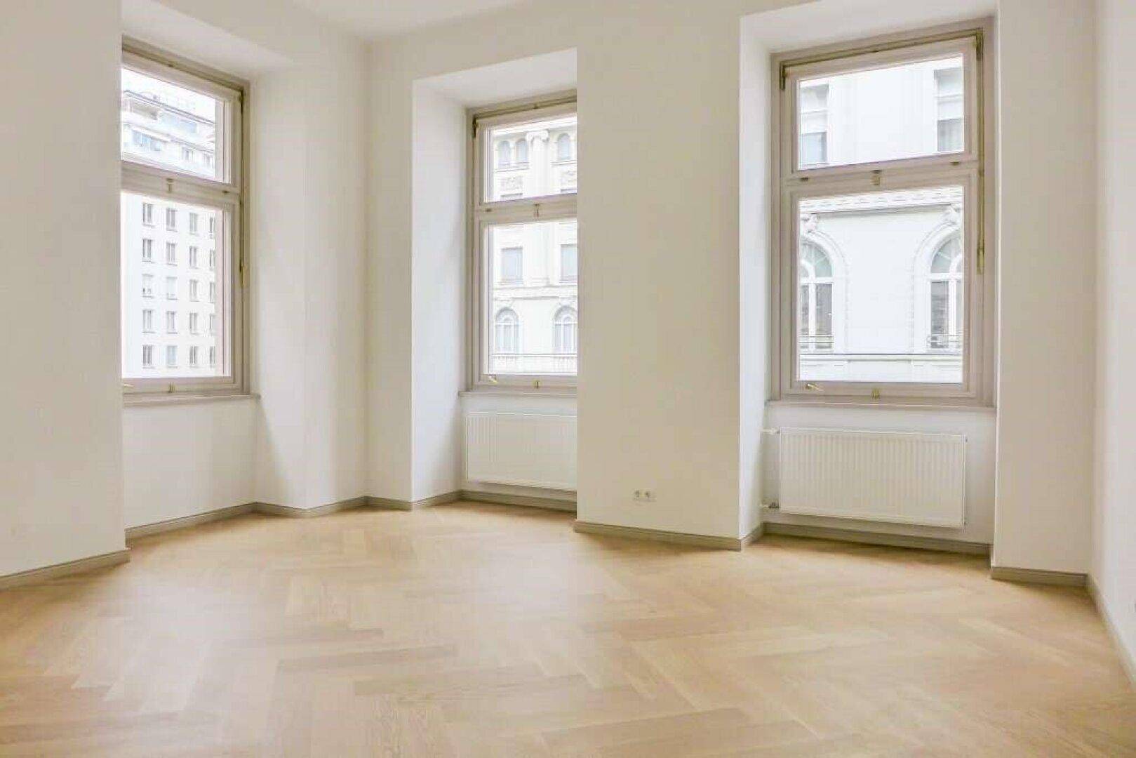 Heller, leerer Raum mit elegantem Fischgrätparkettboden und drei hohen Fenstern, die viel Tageslicht hereinlassen.