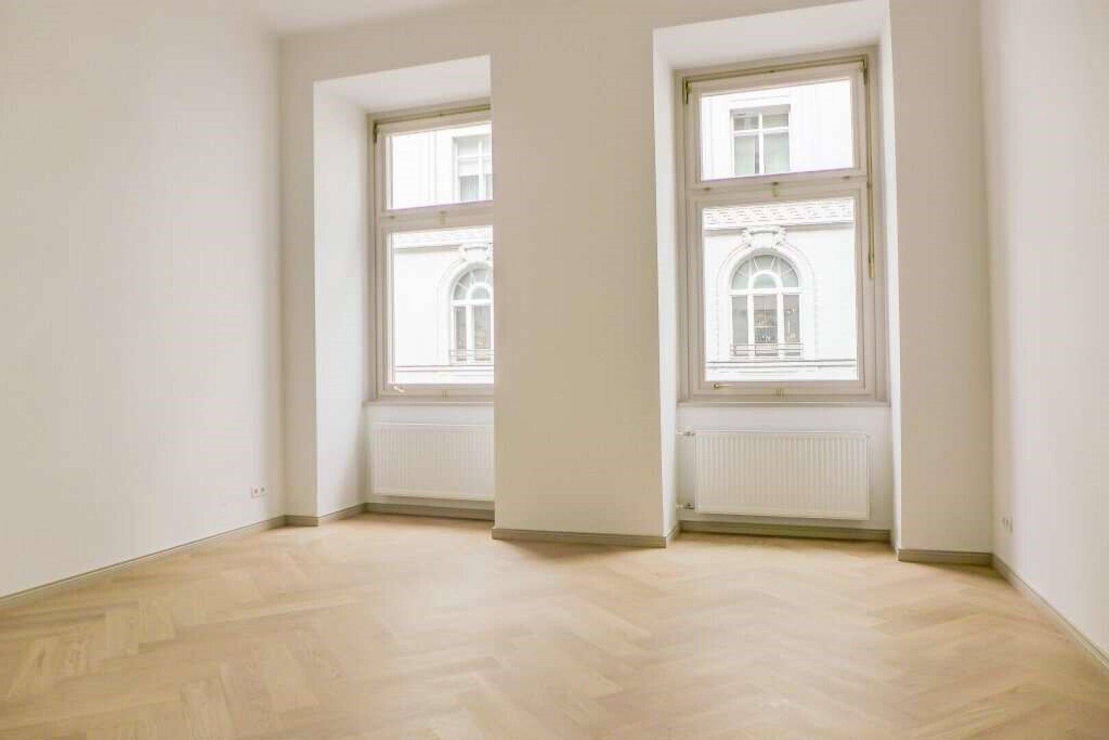 Geräumiger, leerer Raum mit hellem Fischgrätparkett und zwei hohen Fenstern, die den Raum erhellen.