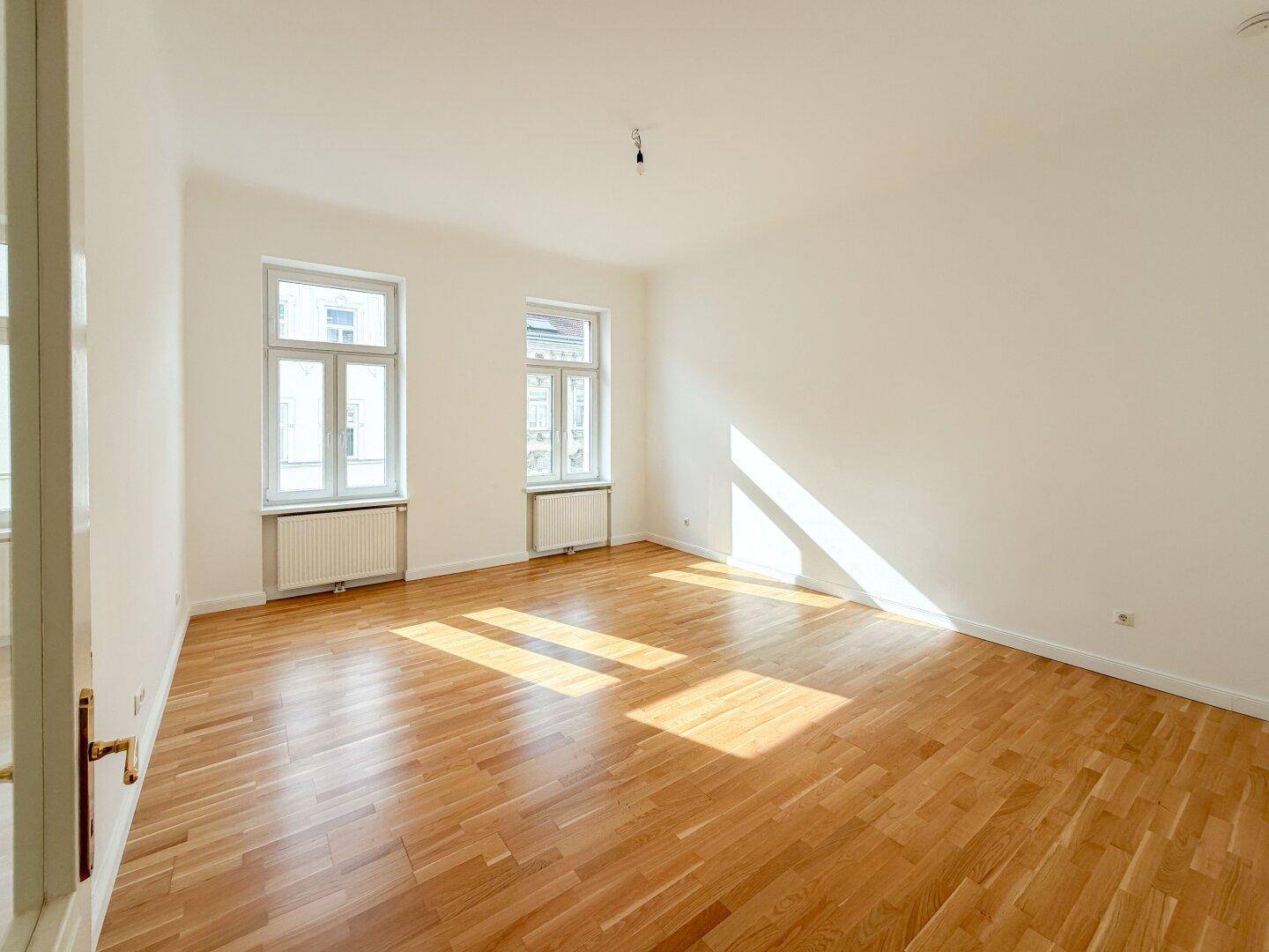 Geräumiges, leeres Wohnzimmer mit Holzboden und viel natürlichem Licht durch die Fenster.