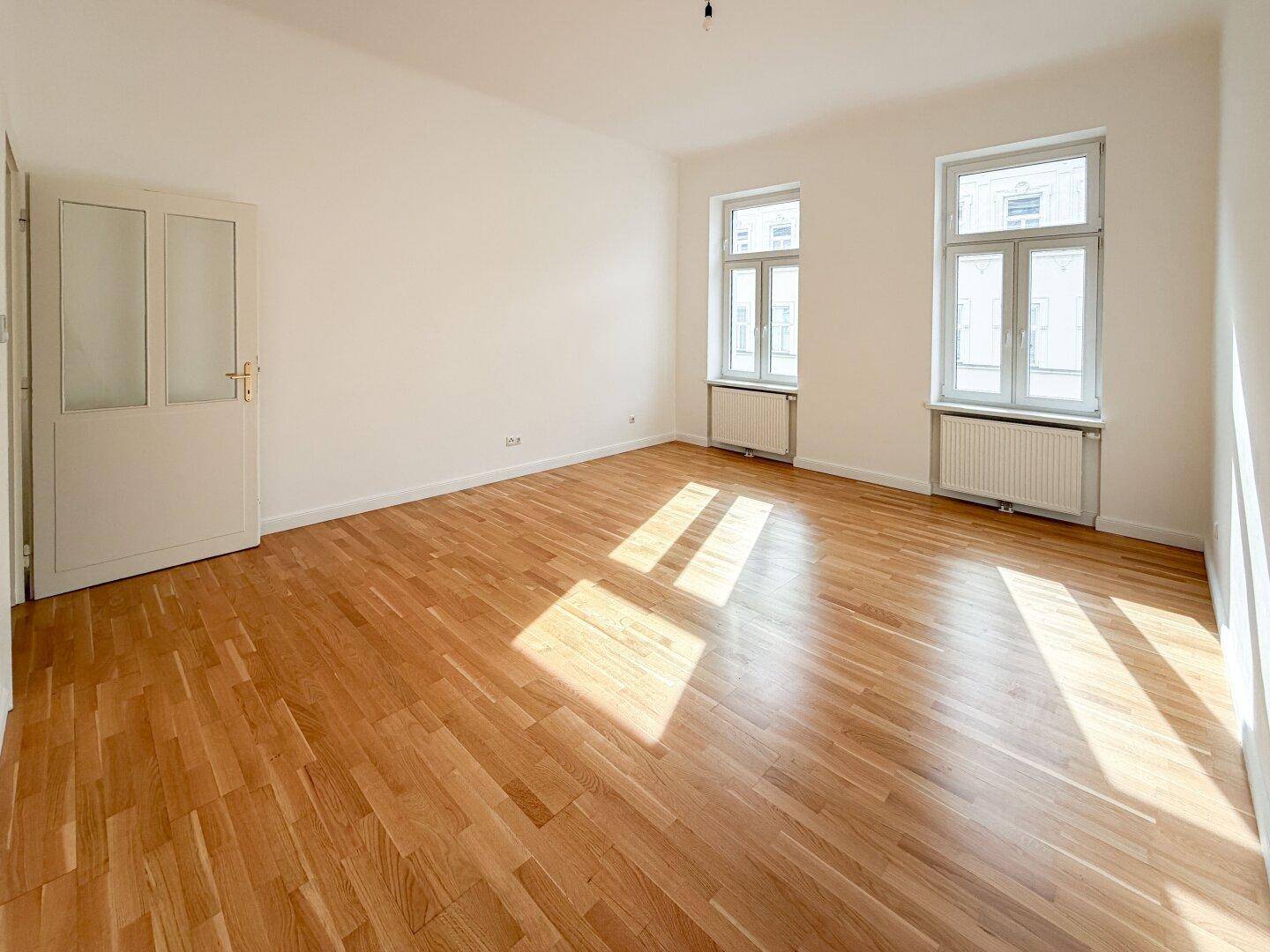 Großes, leeres Wohnzimmer mit Holzboden und zwei Fenstern, die viel Tageslicht hereinlassen.
