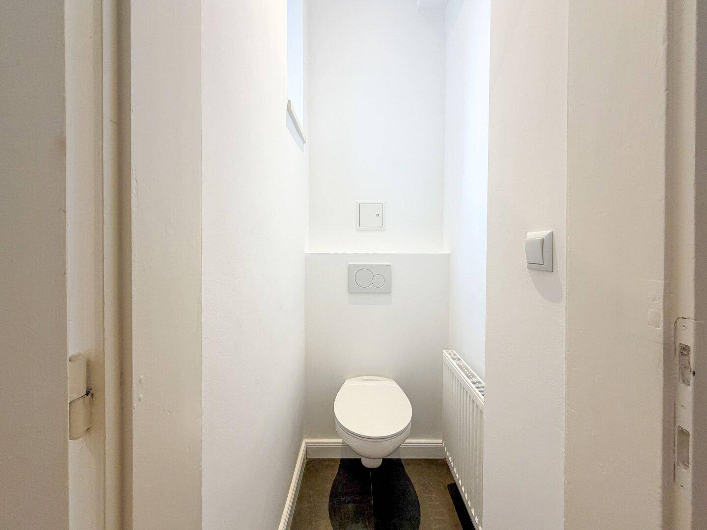 Separates WC mit wandhängender Toilette und kleinem Fenster, in schlichtem, weißem Design.