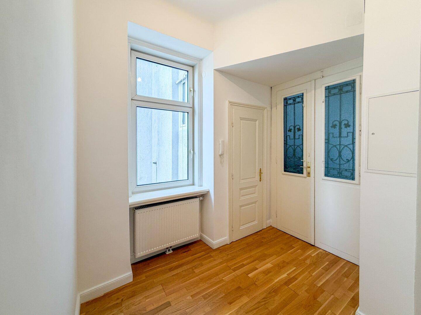 Eingangsbereich mit Holzboden, Fenster und einer Tür mit dekorativem Glas, die zum Flur führt.