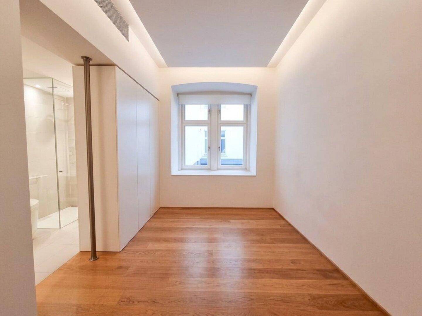 Geräumiges Schlafzimmer mit Holzboden und Fenster, angrenzend an ein modernes Badezimmer mit Glaswand.