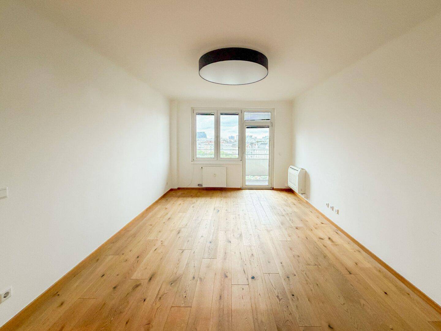 Helles Zimmer mit neuem Holzparkettboden, weißen Wänden und Zugang zu einem kleinen Balkon.