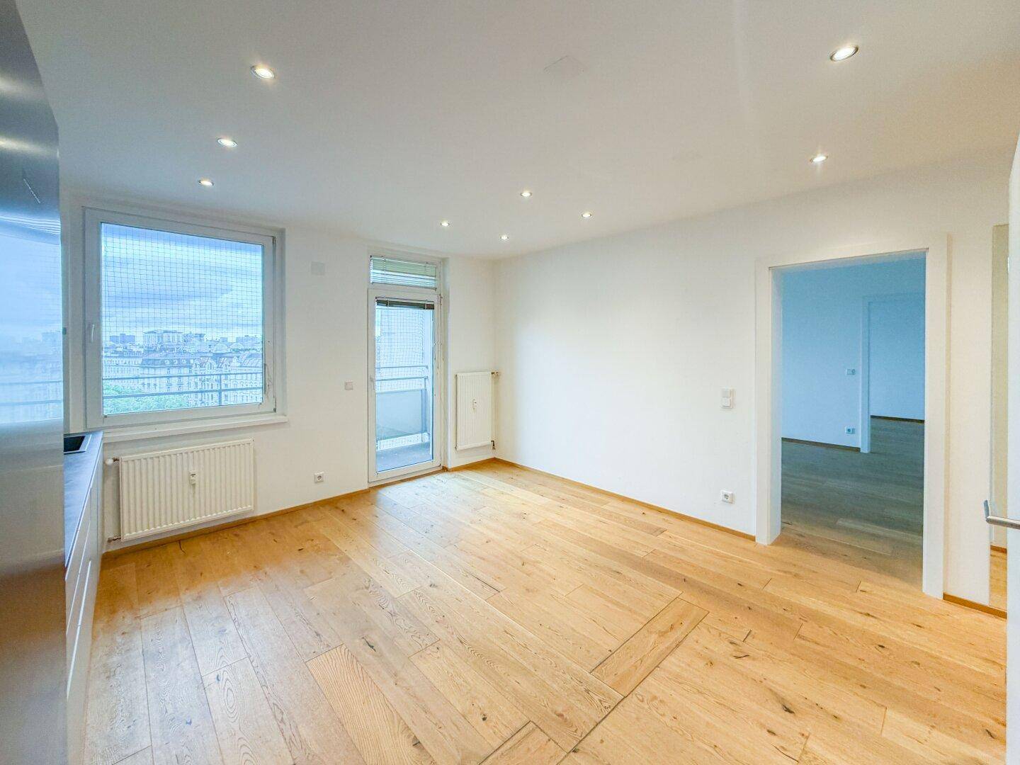 Heller Wohnbereich mit integrierter Küche, Holzparkettboden und direktem Zugang zum Balkon.