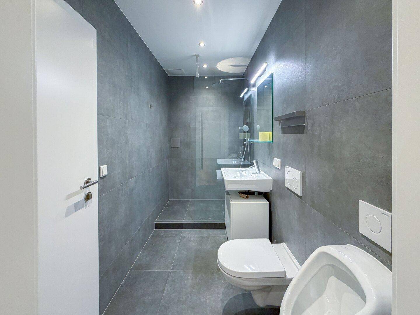 Modernes Badezimmer mit grauen Fliesen, begehbarer Dusche, Wand-WC und Urinal.