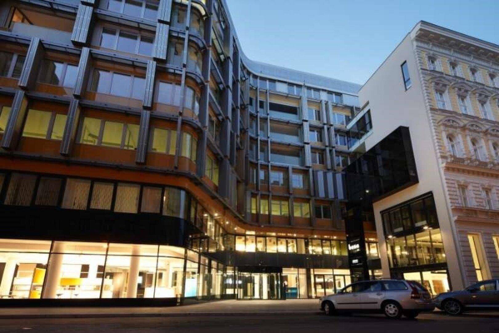 Modernes Geschäftsgebäude mit beleuchteten Innenräumen und angrenzendem Altbau in der Dämmerung.