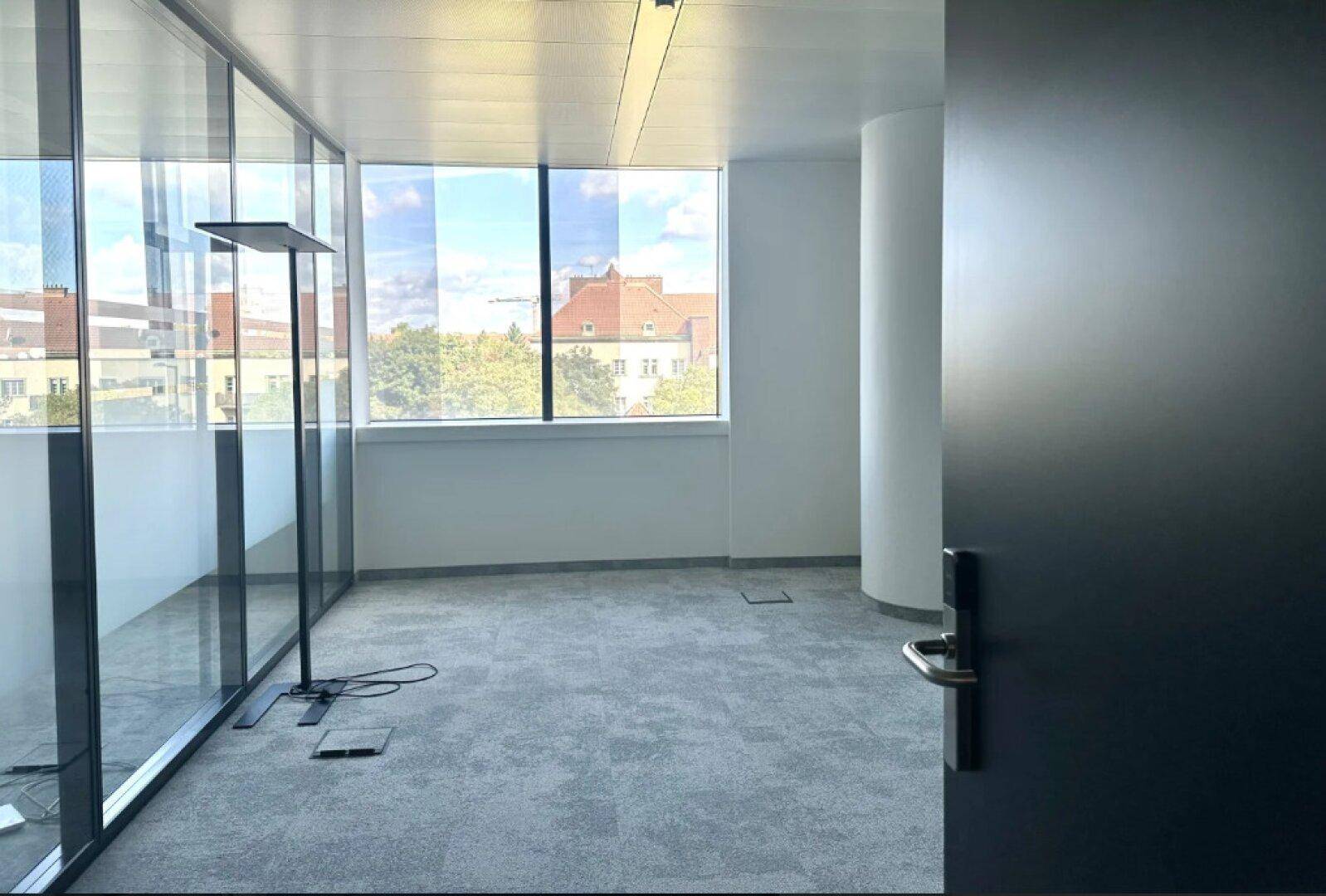 Leeres, helles Büro mit großen Fenstern, Teppichboden und einer modernen Stehleuchte.