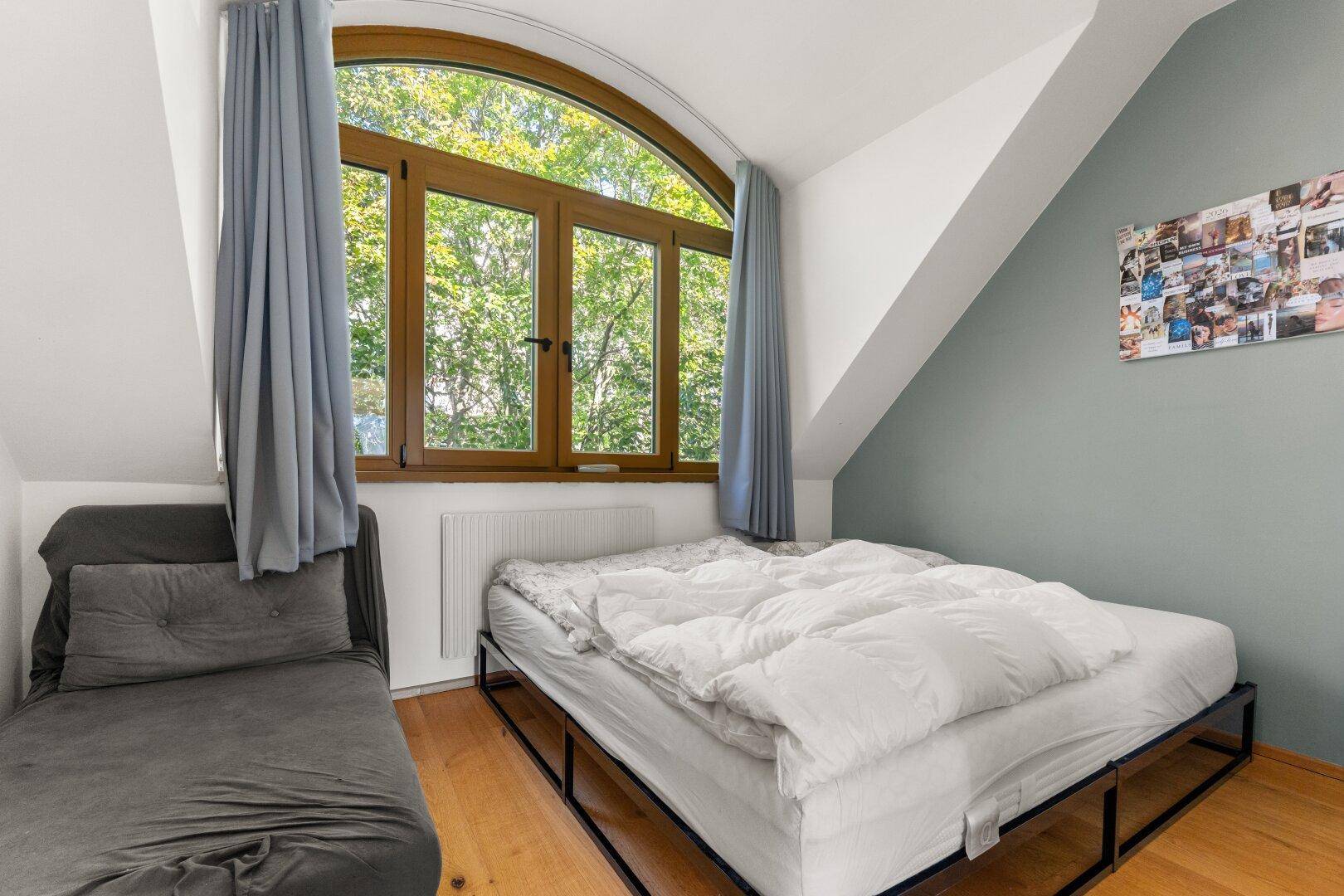 Schlafzimmer mit Bett, Tagesliege und großem Bogenfenster mit Blick ins Grüne.
