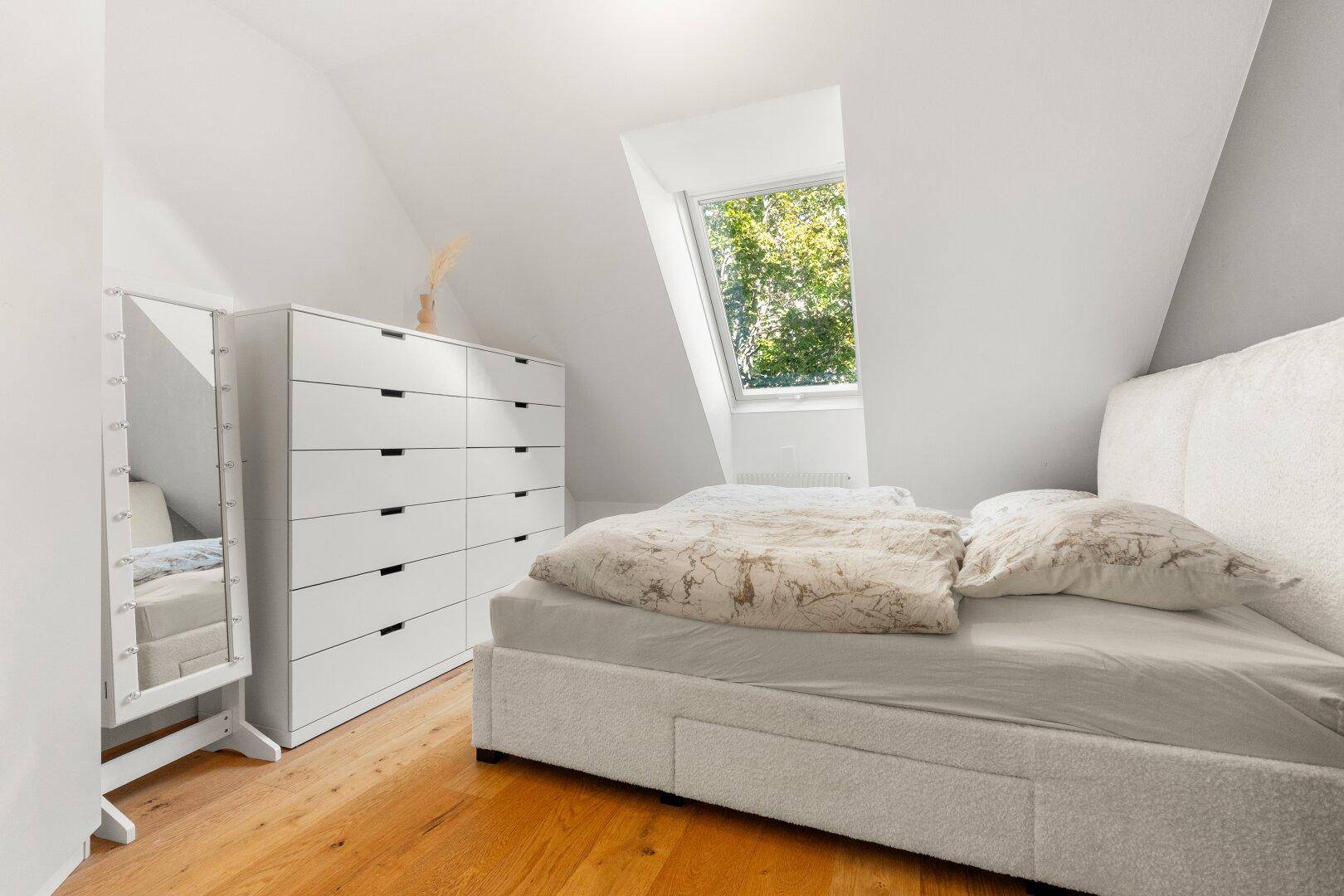 Schlafzimmer mit Bett, Kommode, Spiegel und Dachfenster im hellen Dachgeschoss.
