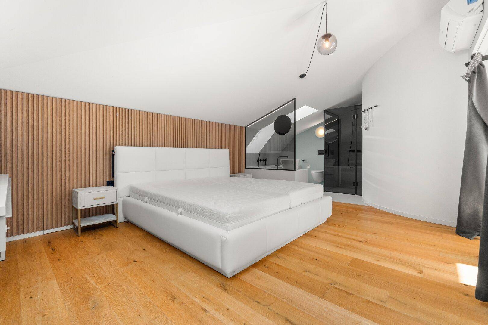 Hauptschlafzimmer mit großem Bett, Holzwand und offenem Badbereich mit Glasabtrennung.