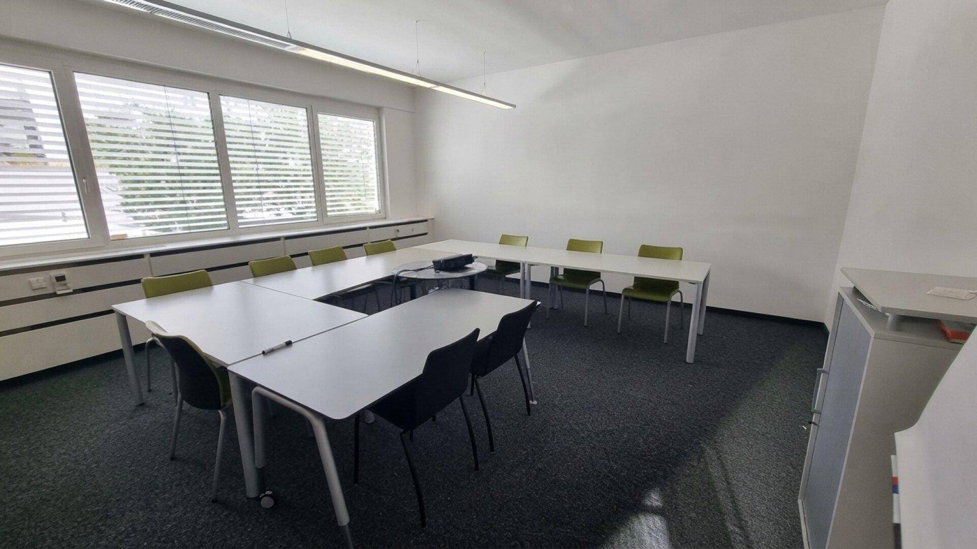 Heller Konferenzraum mit U-förmiger Tischordnung, modernen Stühlen und großen Fenstern für viel Tageslicht.