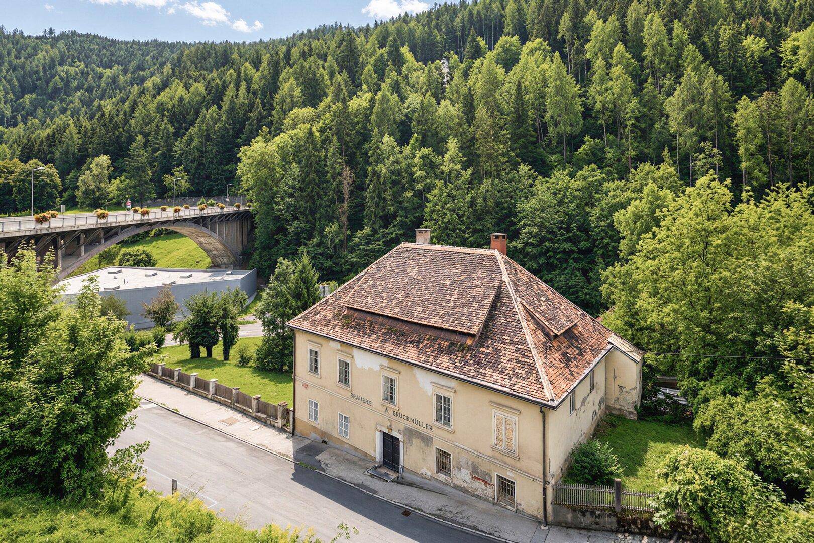 Historisches Anwesen mit Ziegeldach und Brücke im Hintergrund, eingebettet in grüne Natur.