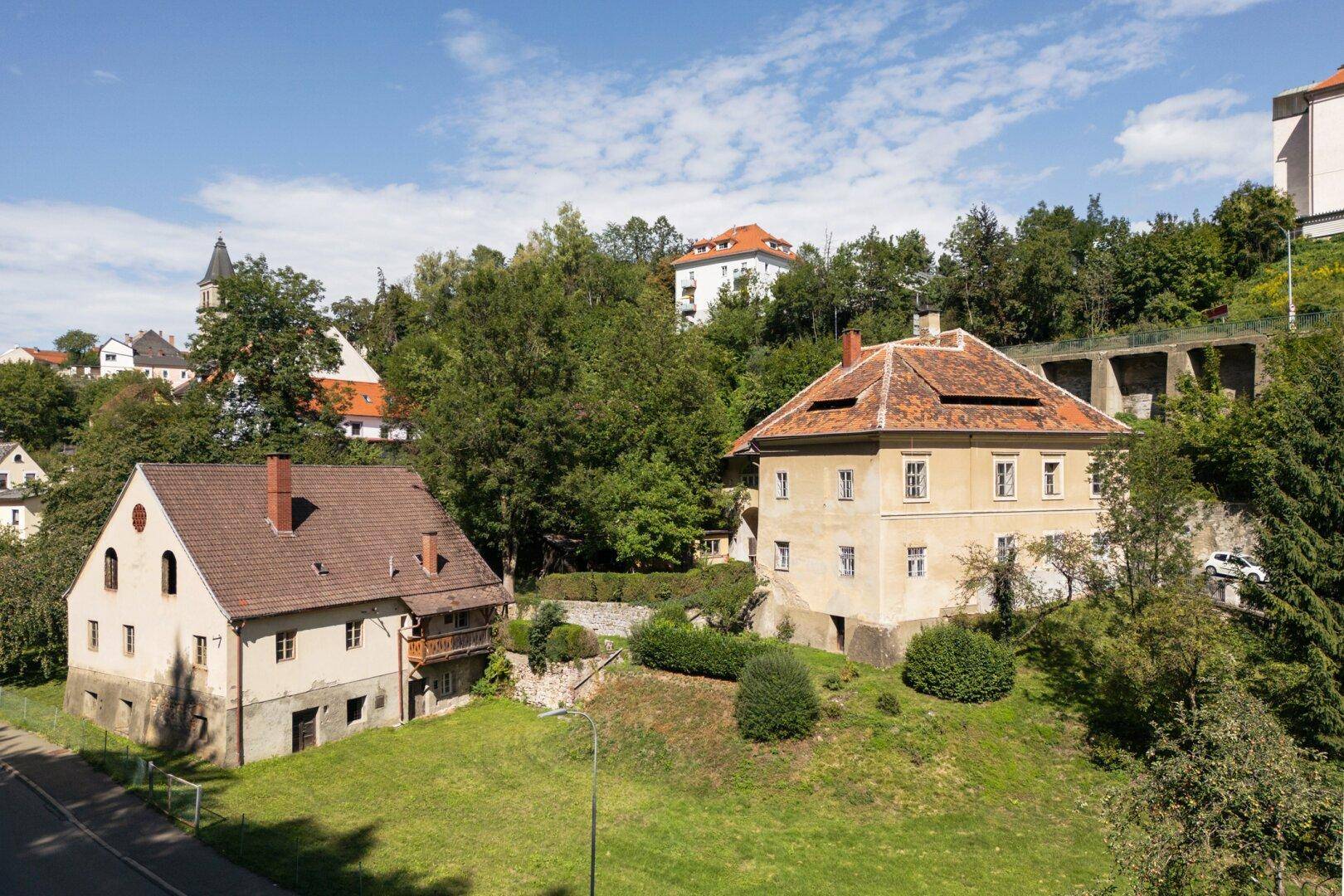 Historisches Anwesen mit zwei Gebäuden, umgeben von grünen Hügeln und Bäumen.