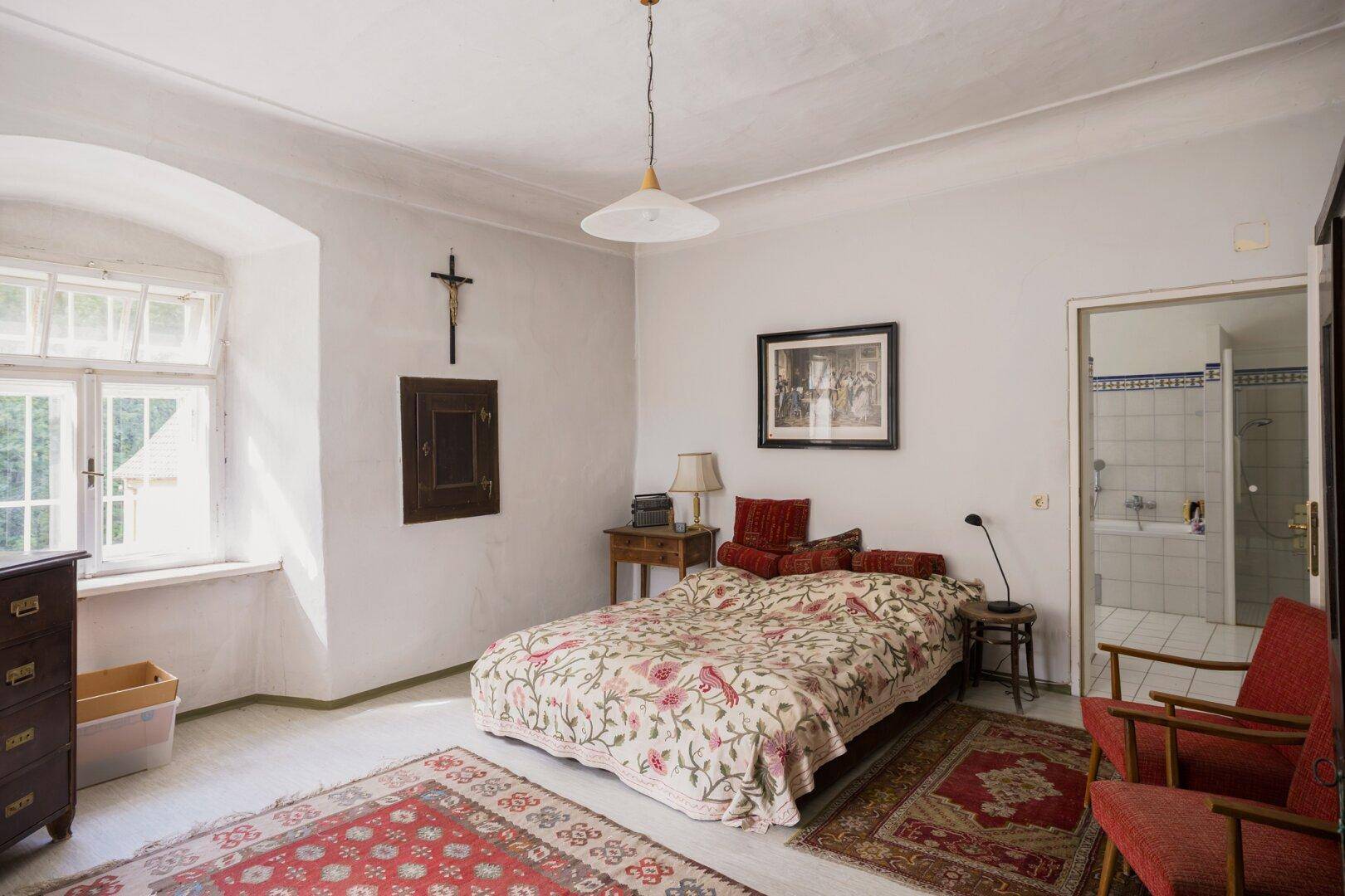 Helles Schlafzimmer mit traditionellem Bett, Teppich und Blick ins Grüne durch das Fenster.