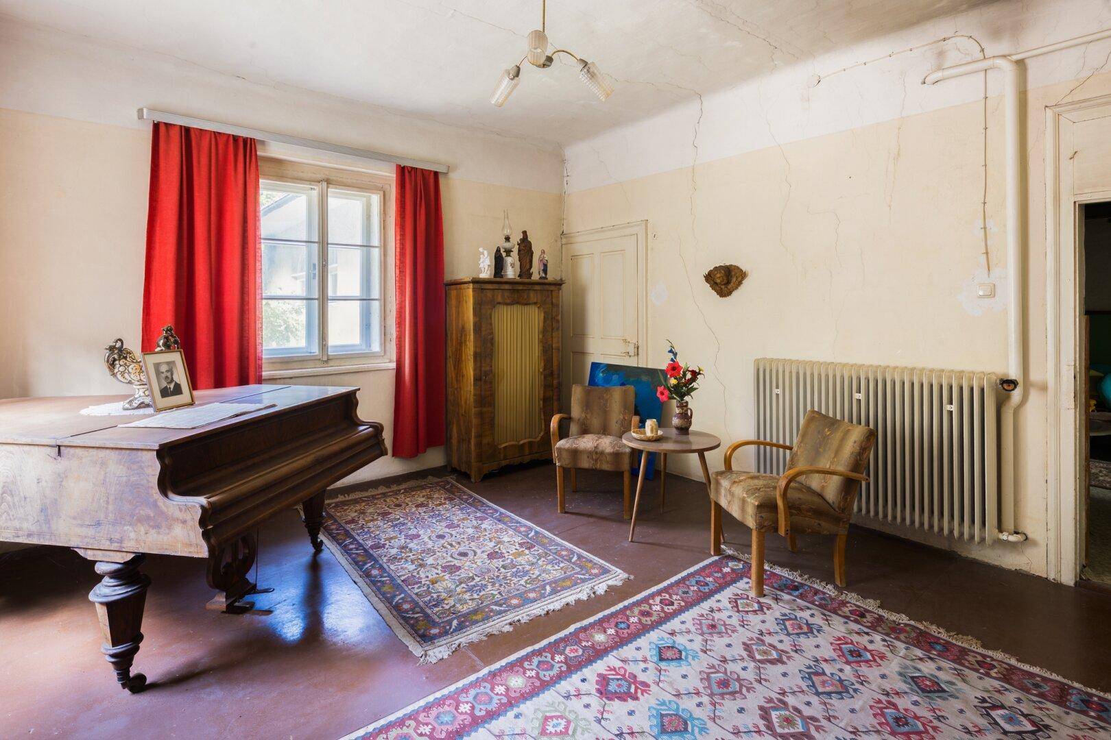 Zimmer mit Klavier, traditionellen Möbeln, Teppichen und großem Heizkörper.