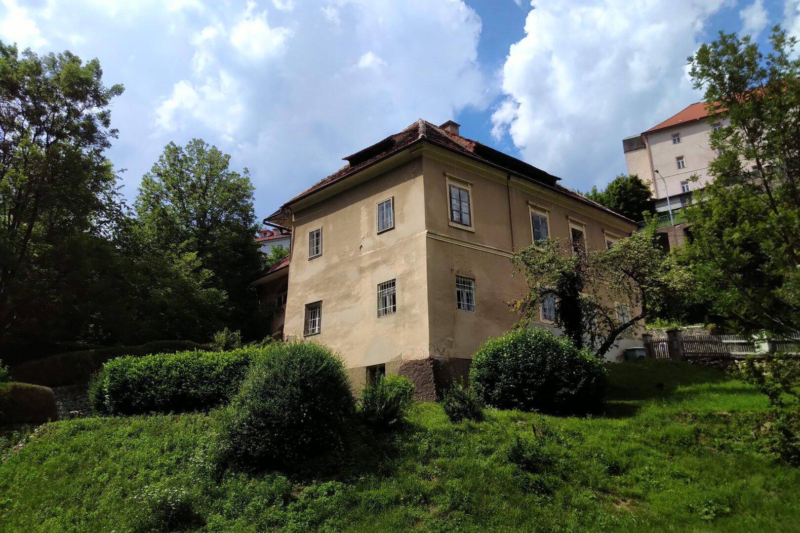 Historisches Gebäude mit gelber Fassade und Ziegeldach, umgeben von gepflegtem Grün.